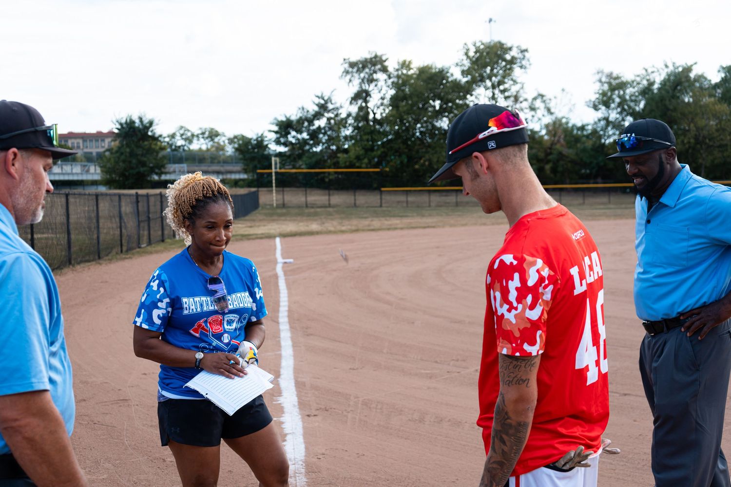 Four people conversing on a baseball field, including a woman in a blue jersey holding papers and a man in a red uniform.