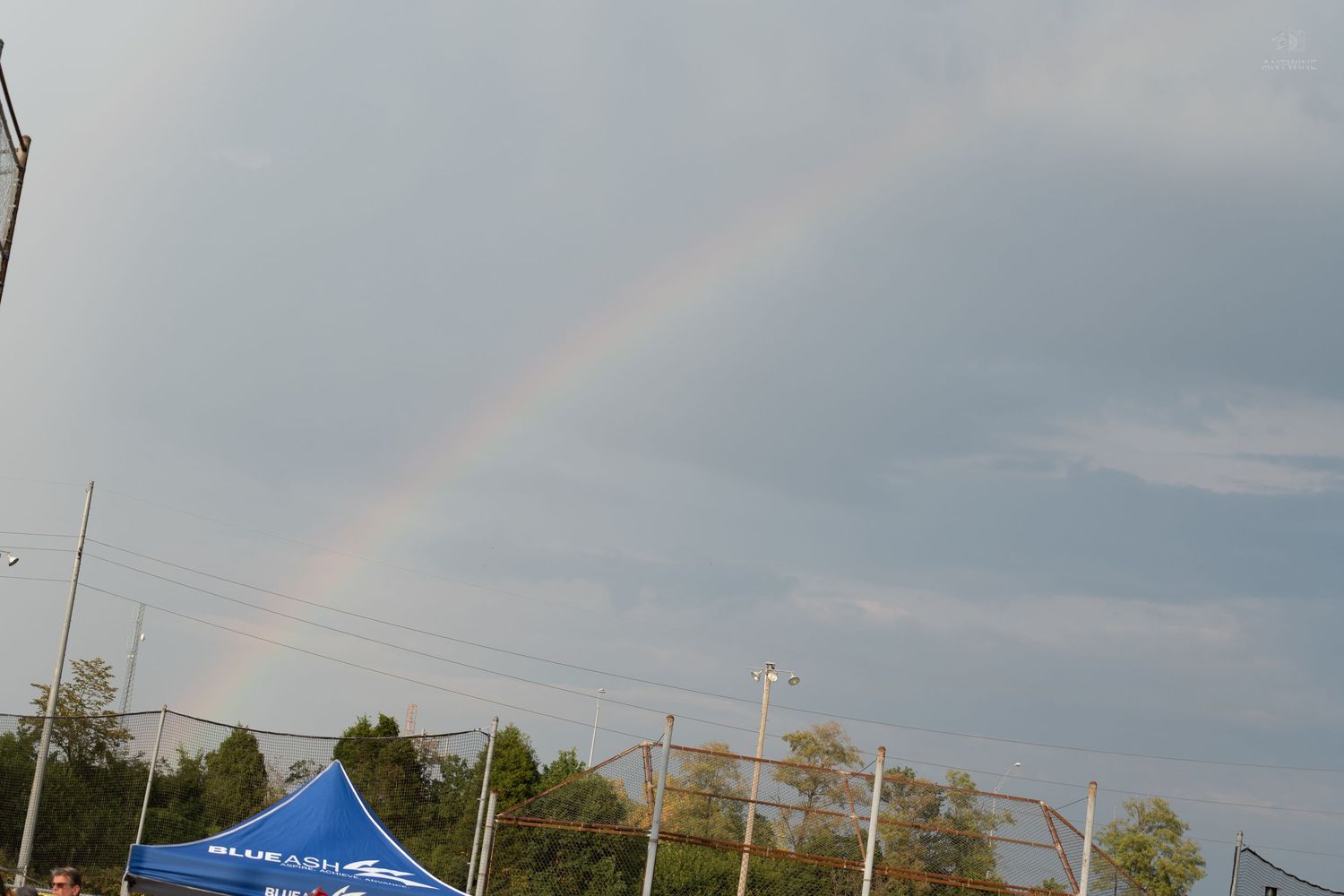 A vibrant rainbow arcs across a cloudy sky above trees and a blue event tent at an outdoor venue.