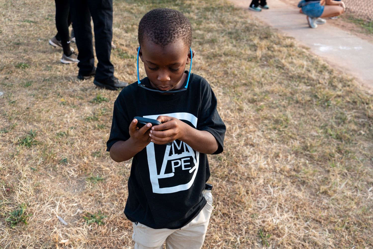 Young Black boy looking down at a small object while standing on dry grass outdoors.
