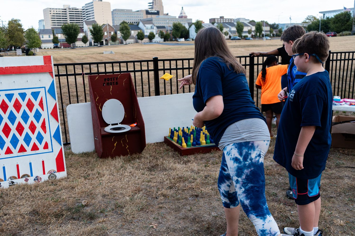 Children and adult playing carnival games outdoors at a community fair event near a fenced field.