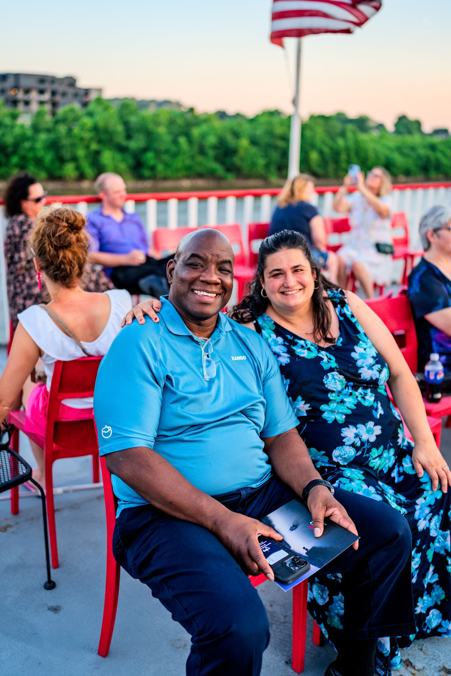 Smiling couple poses together on a riverboat cruise at sunset with American flag and guests in background.