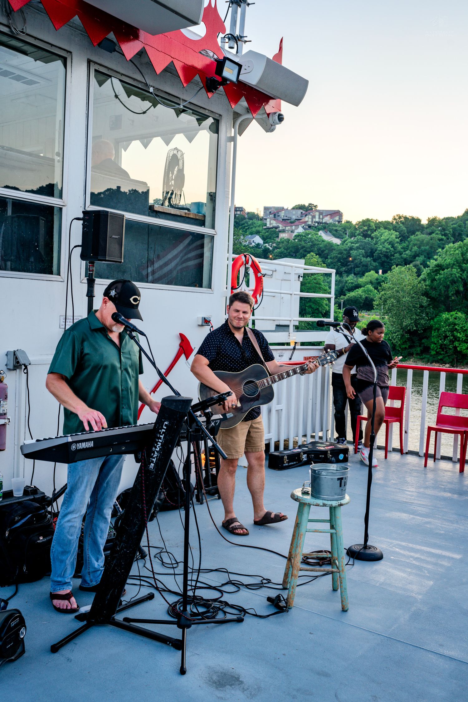Live band performing on a riverboat deck with guitars and keyboard at sunset.