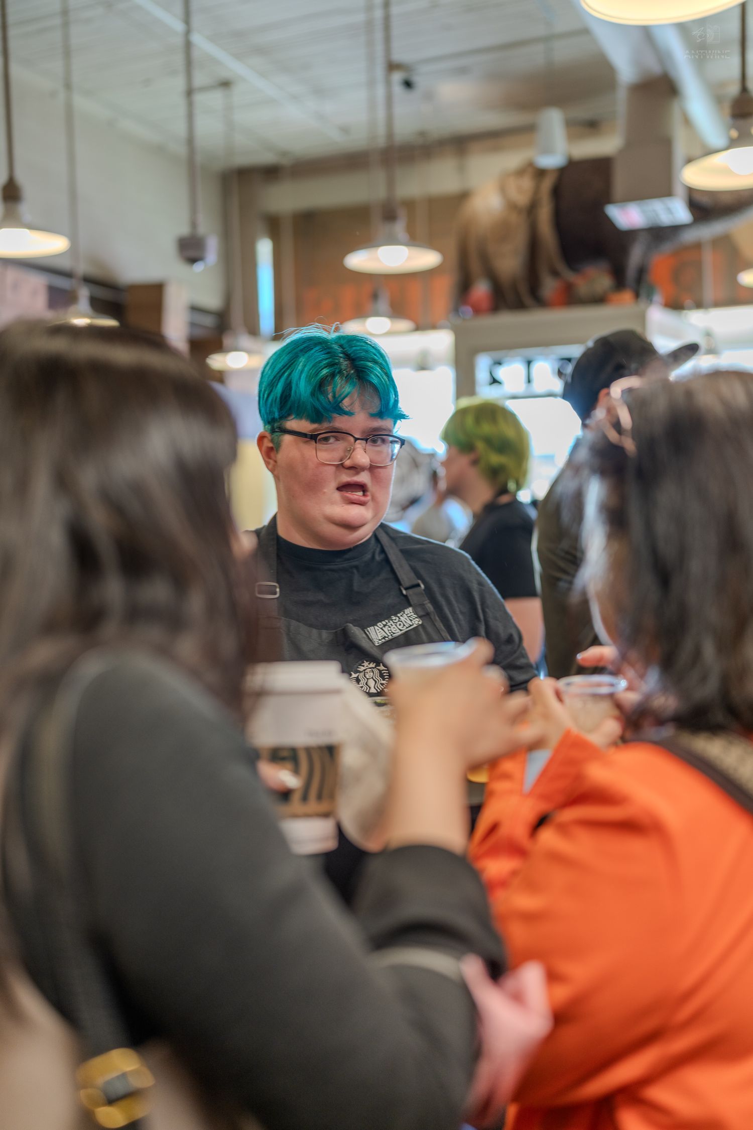 Starbucks barista with teal hair serving customers in a busy coffee shop.