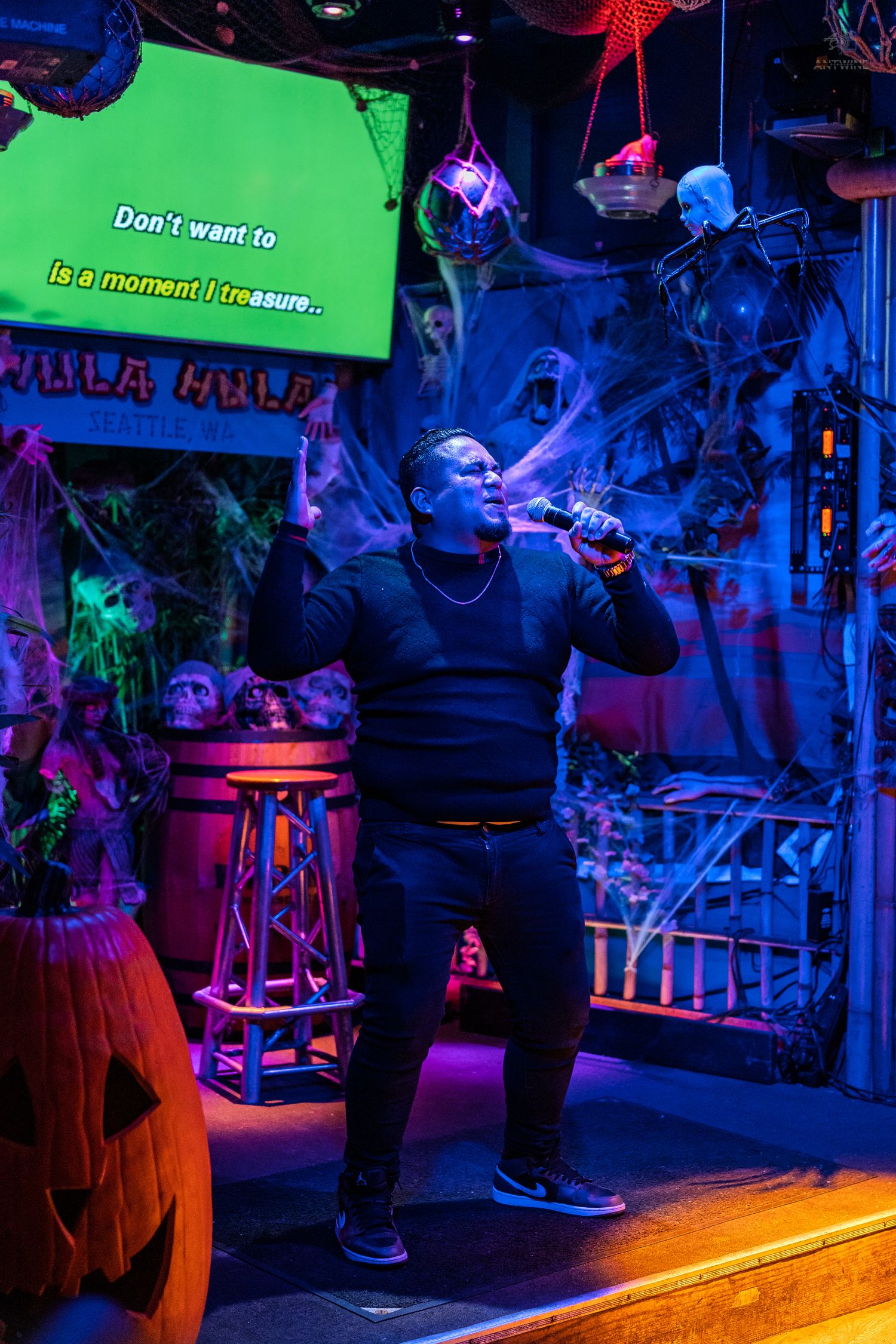 Man singing karaoke at Halloween-decorated Hula Hula bar in Seattle under colorful neon lights.