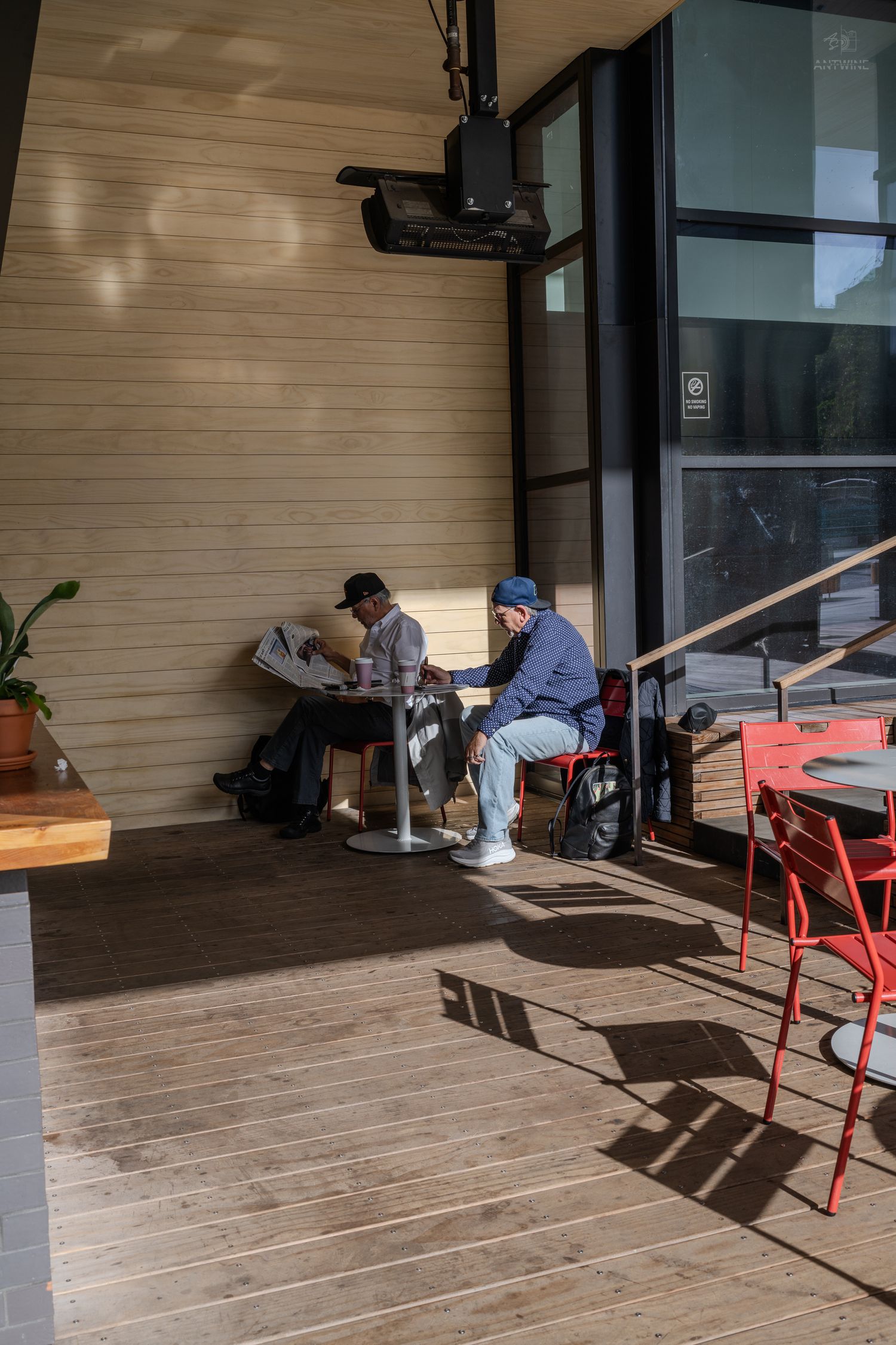 Two people sitting at outdoor cafe tables reading in warm sunlight on a wooden deck patio.