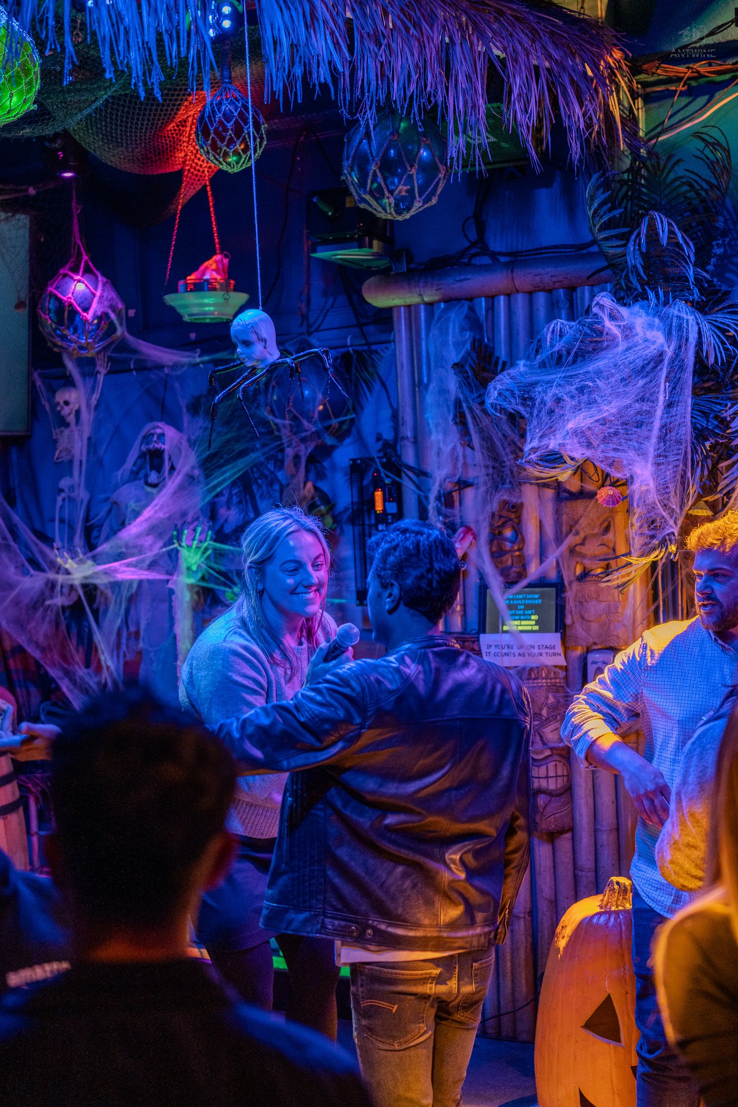 Halloween-themed bar with neon lights, fake spiderwebs, and guests socializing under tiki decorations.