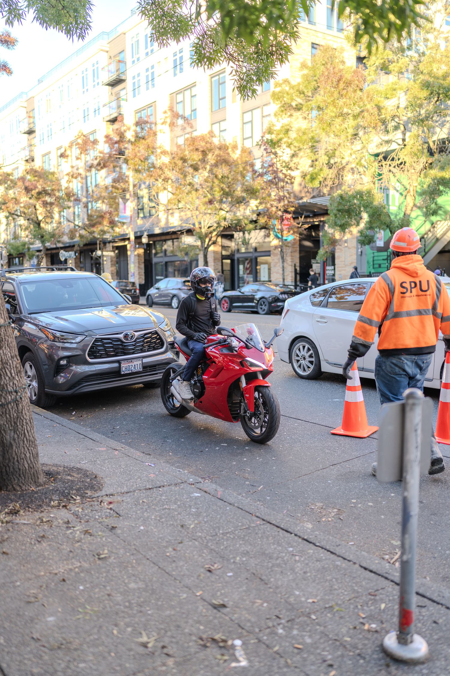 Red motorcycle rider navigating urban street traffic past an SPU worker in orange safety vest with traffic cone.
