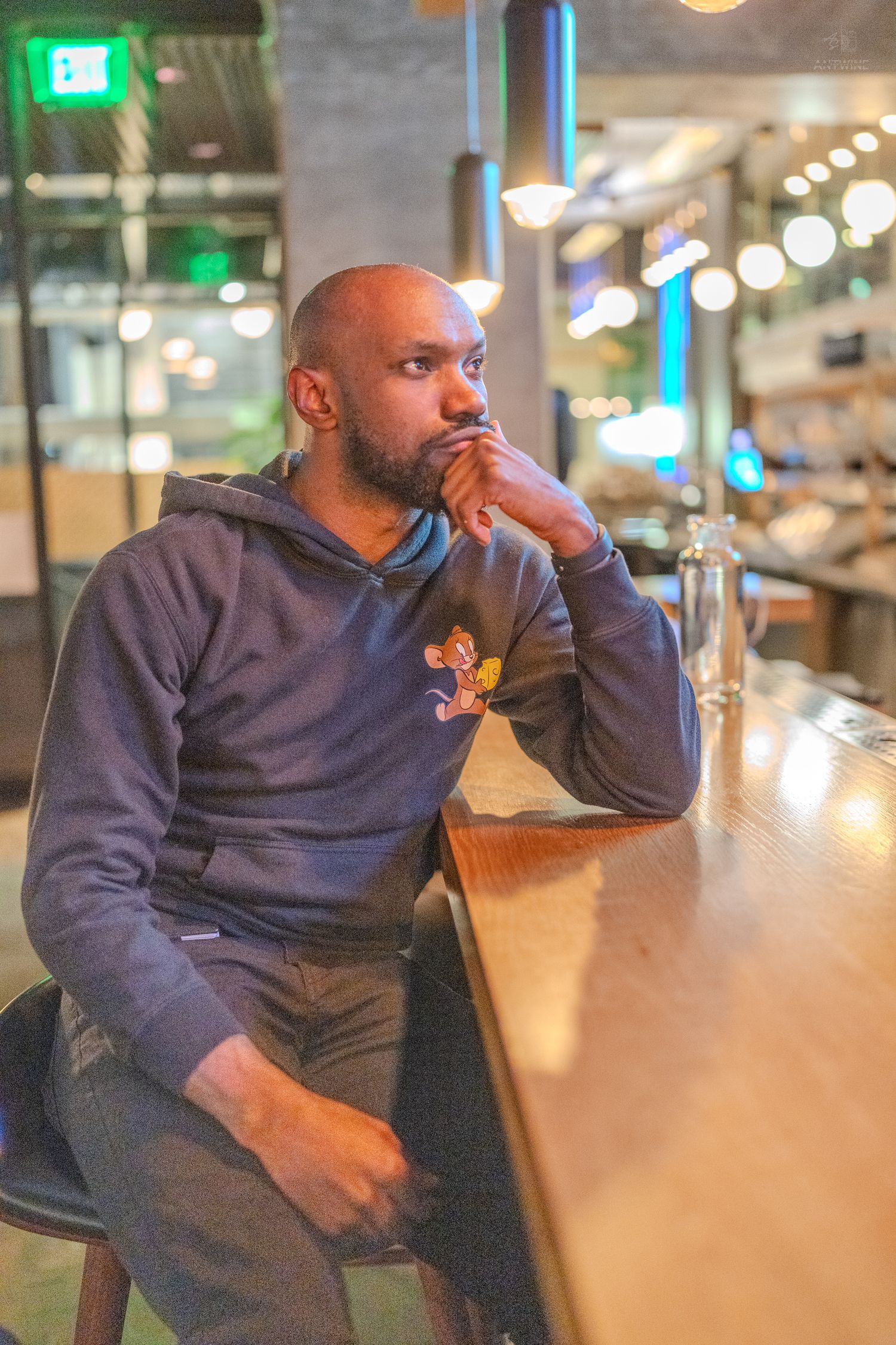 A thoughtful bald man with a beard sits at a bar counter in a modern restaurant.
