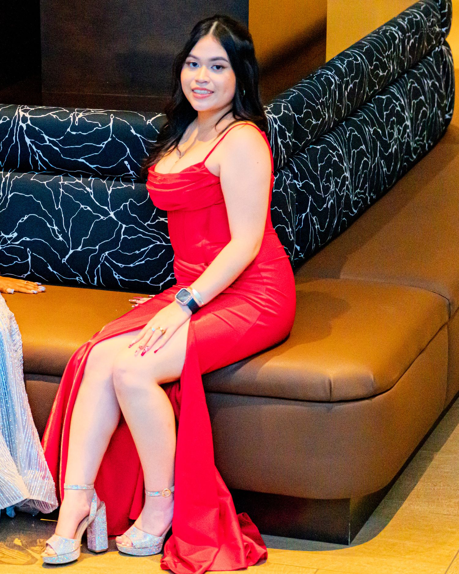 Young woman posing in a stunning red satin mermaid gown with a thigh-high slit, seated on a leather sofa.