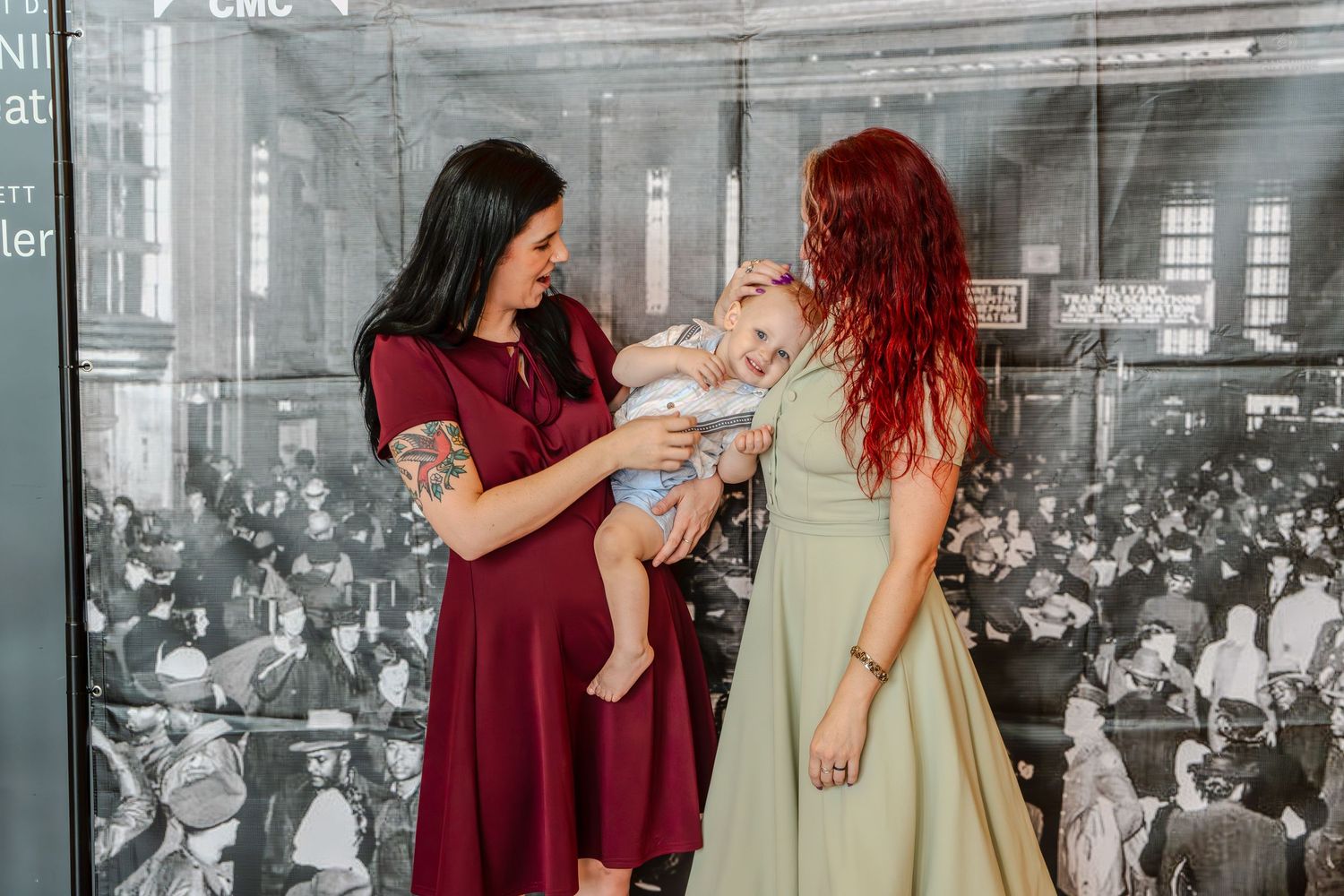 Two women in elegant dresses lovingly interact with a baby at a museum exhibit with a historic black-and-white photo backdrop.