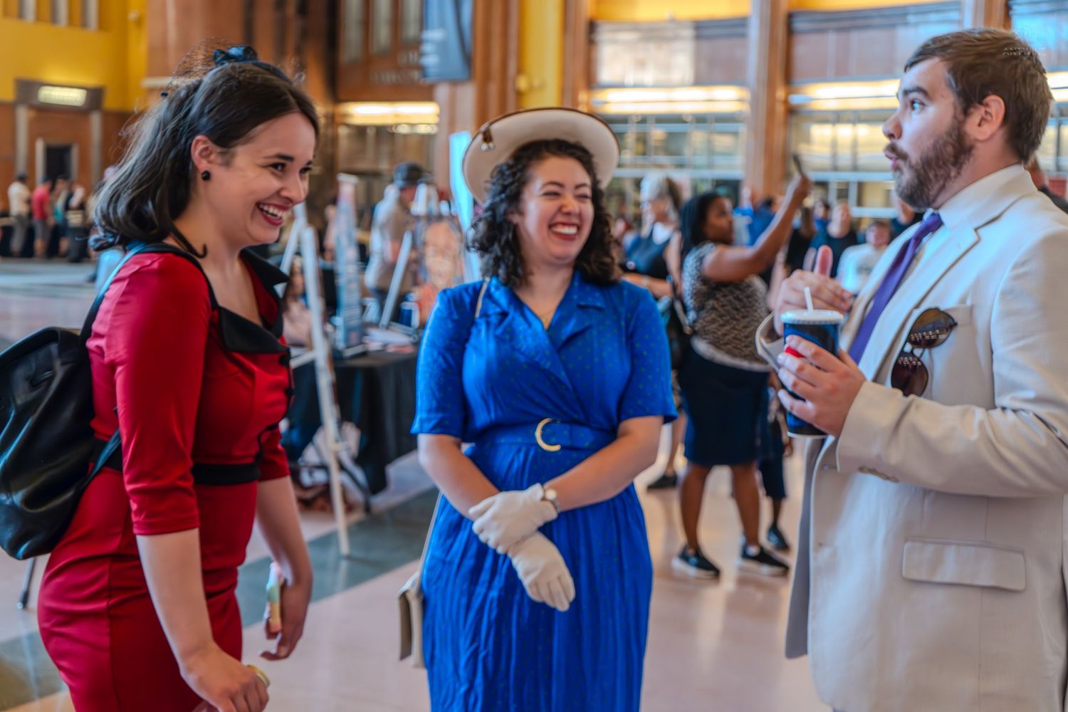 Three people in vintage 1940s attire chat and laugh at an indoor social event, wearing red, blue, and cream outfits.