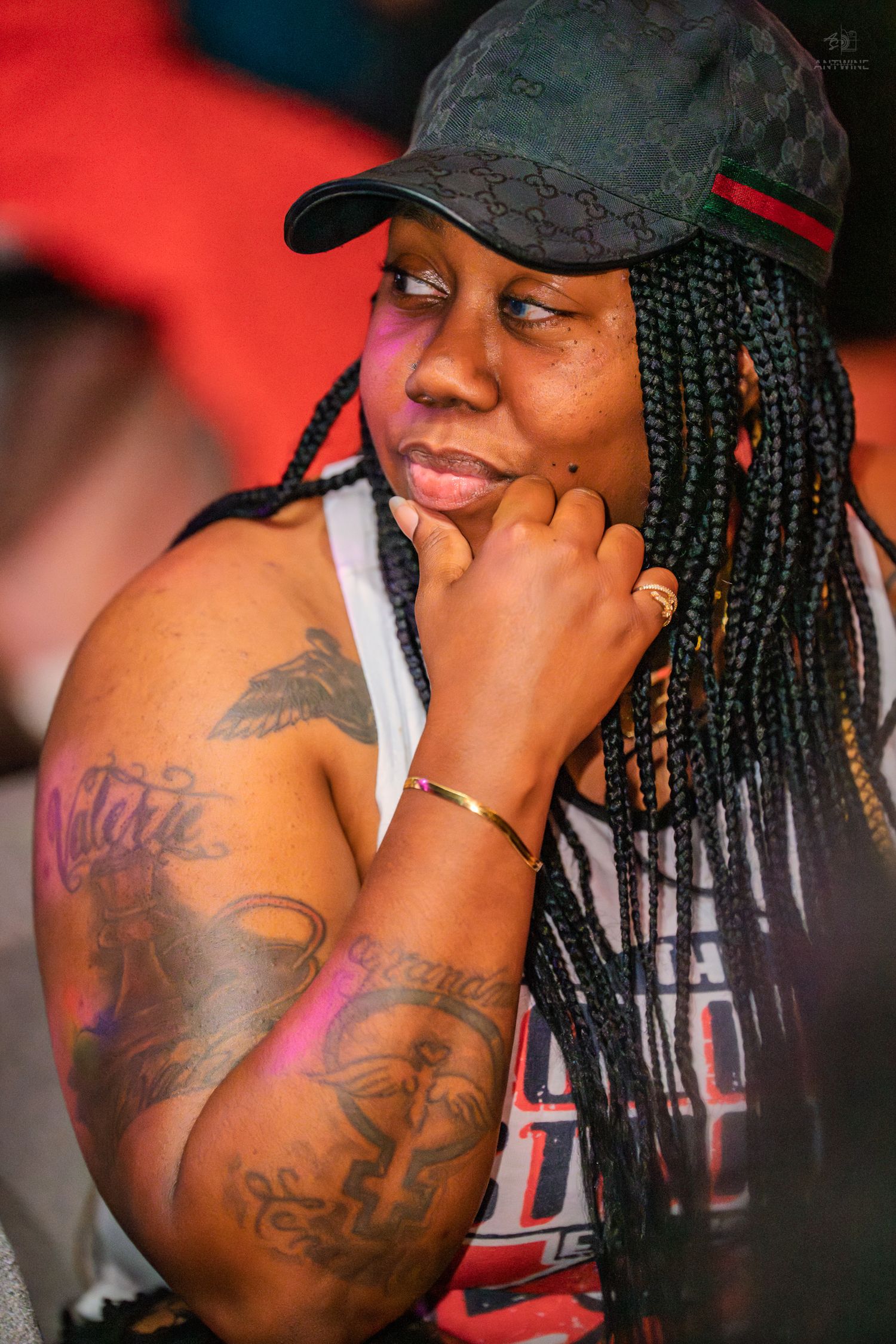 Woman with braids, cap, and tattoos resting chin on hand thoughtfully at an outdoor event.