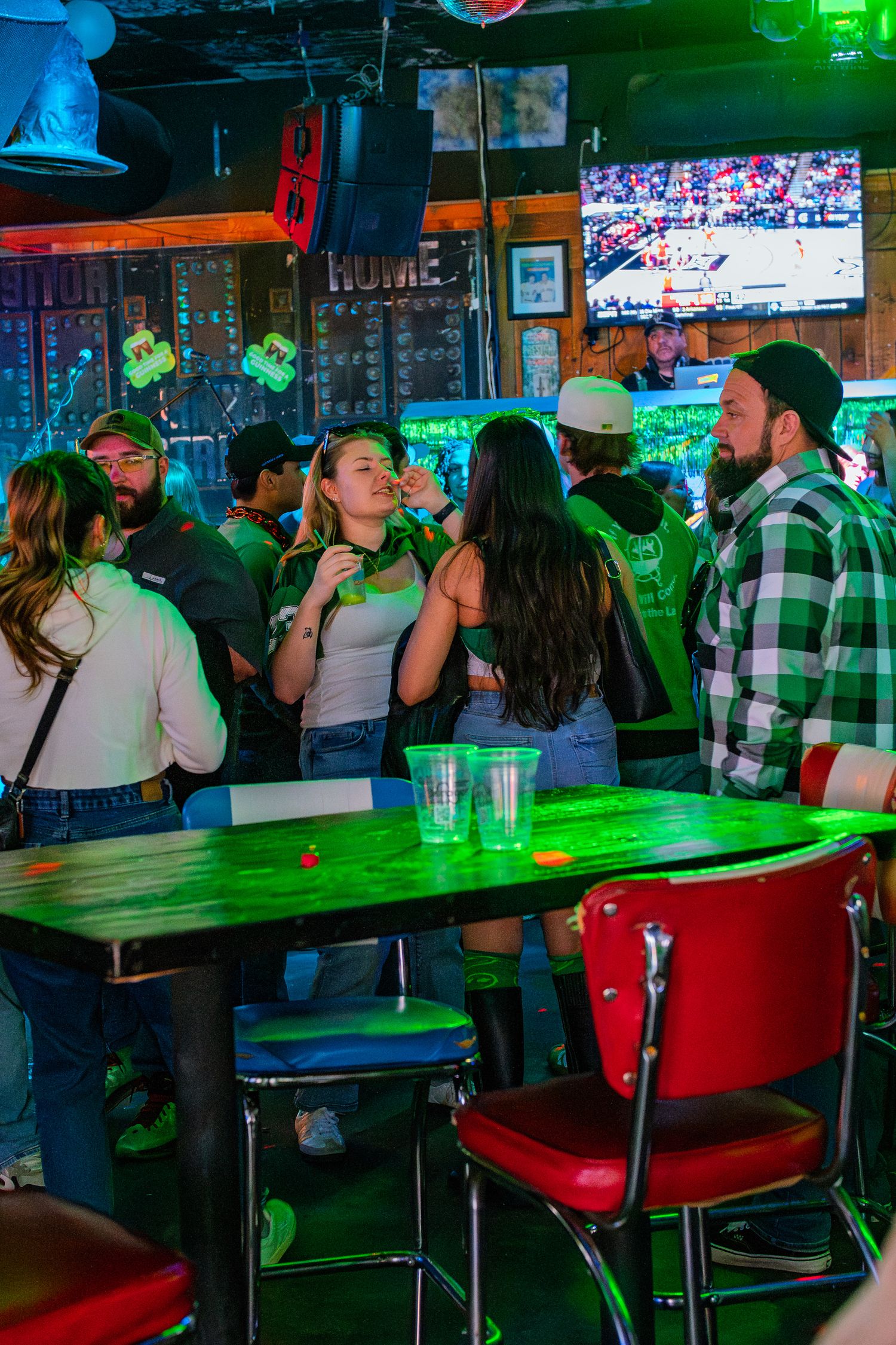Crowded bar with green lighting, people socializing and drinking on St. Patrick's Day near a green table.