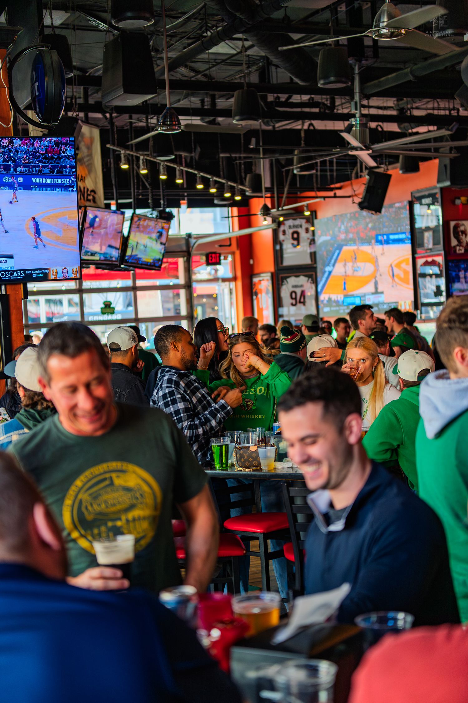Crowded sports bar with fans in green St. Patrick's Day attire watching multiple TVs during a lively celebration.