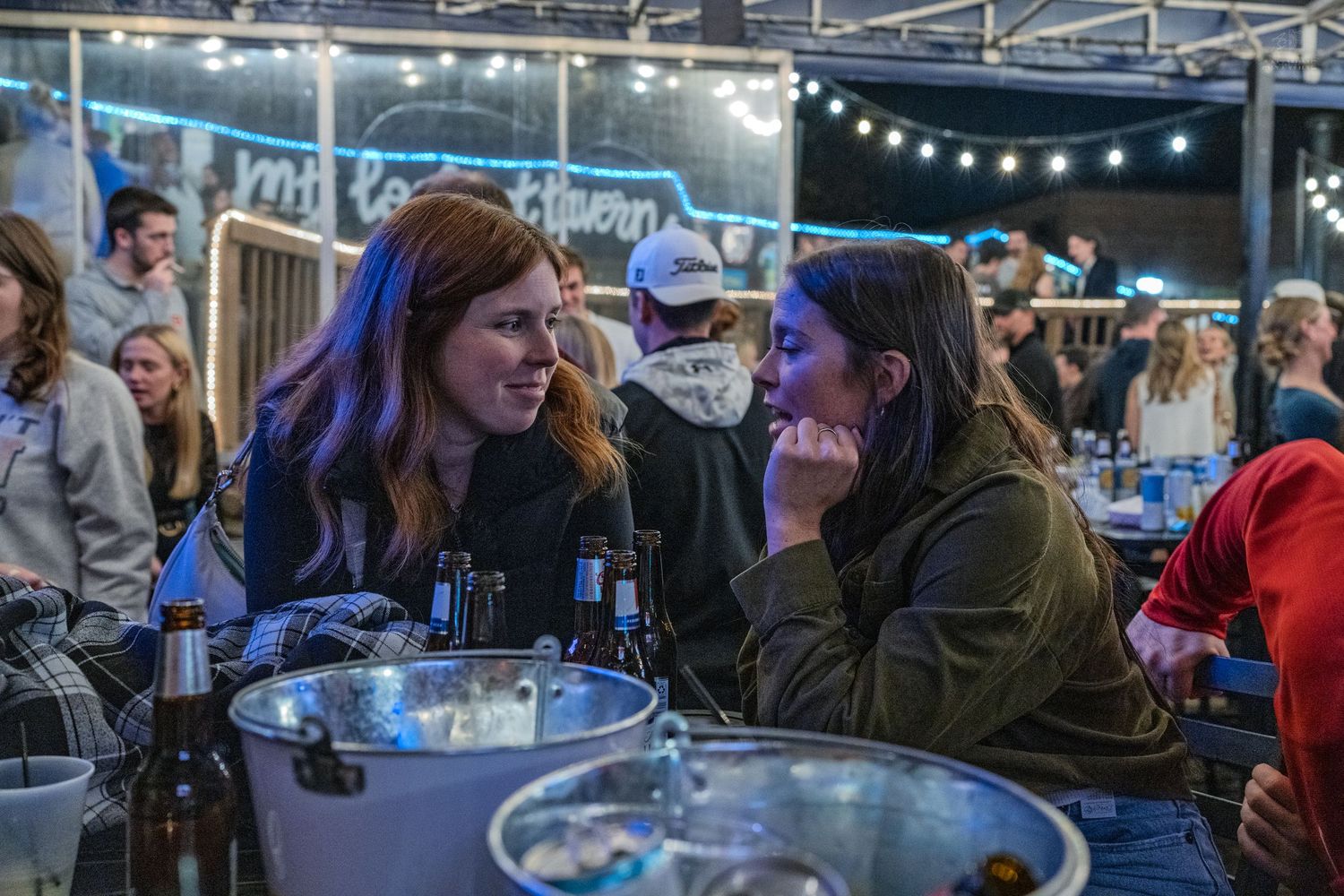 Two women engaged in conversation at a crowded bar with beer bottles on the table.