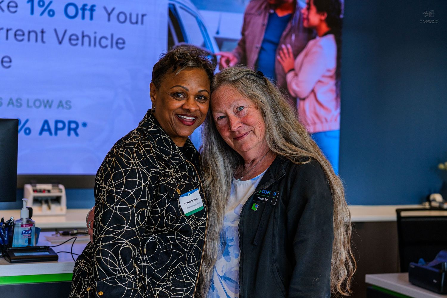 Two women smiling and posing together at a car dealership event with promotional APR signage in the background.