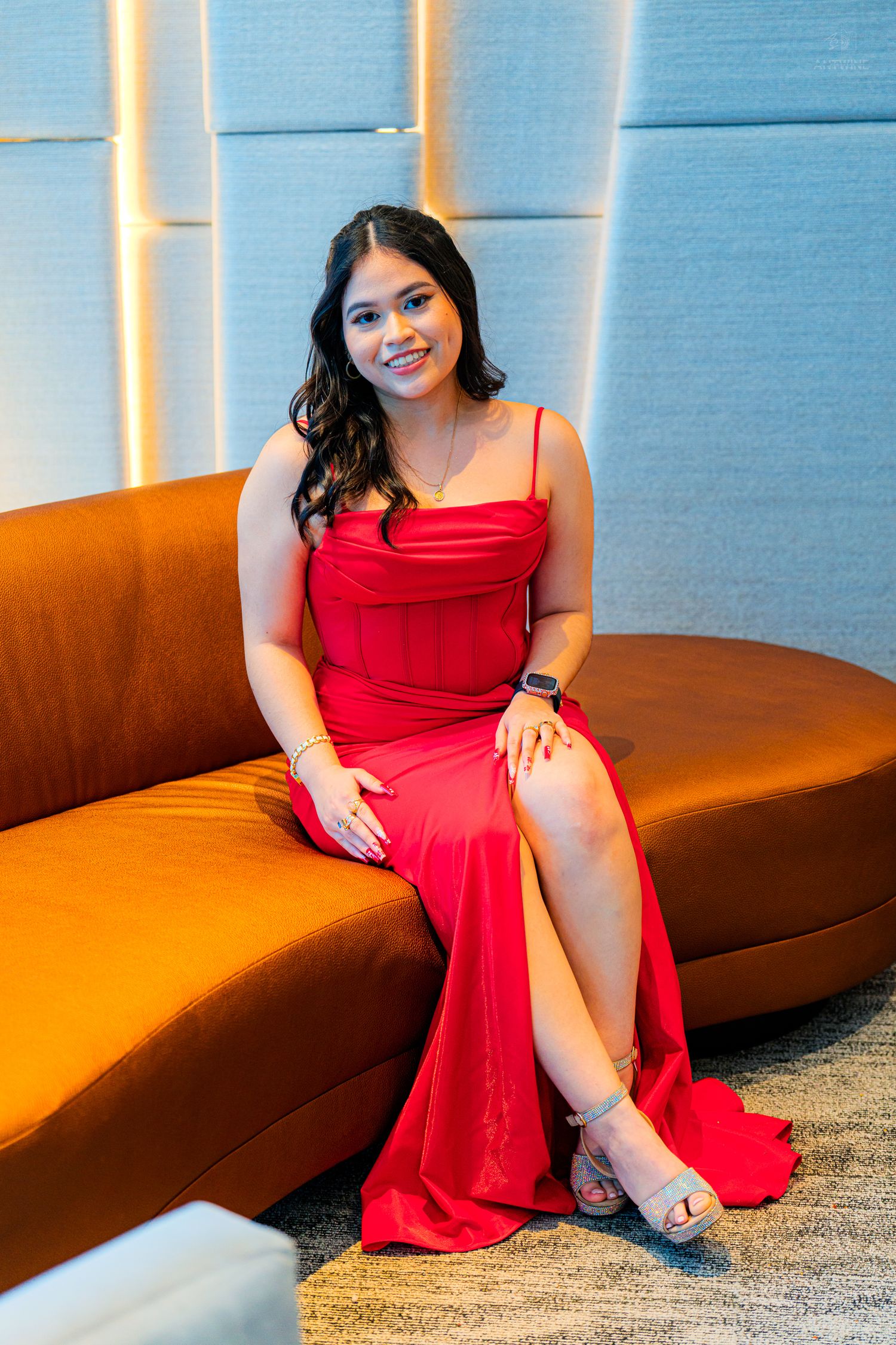Woman in elegant red slit gown seated on curved orange sofa in upscale interior.