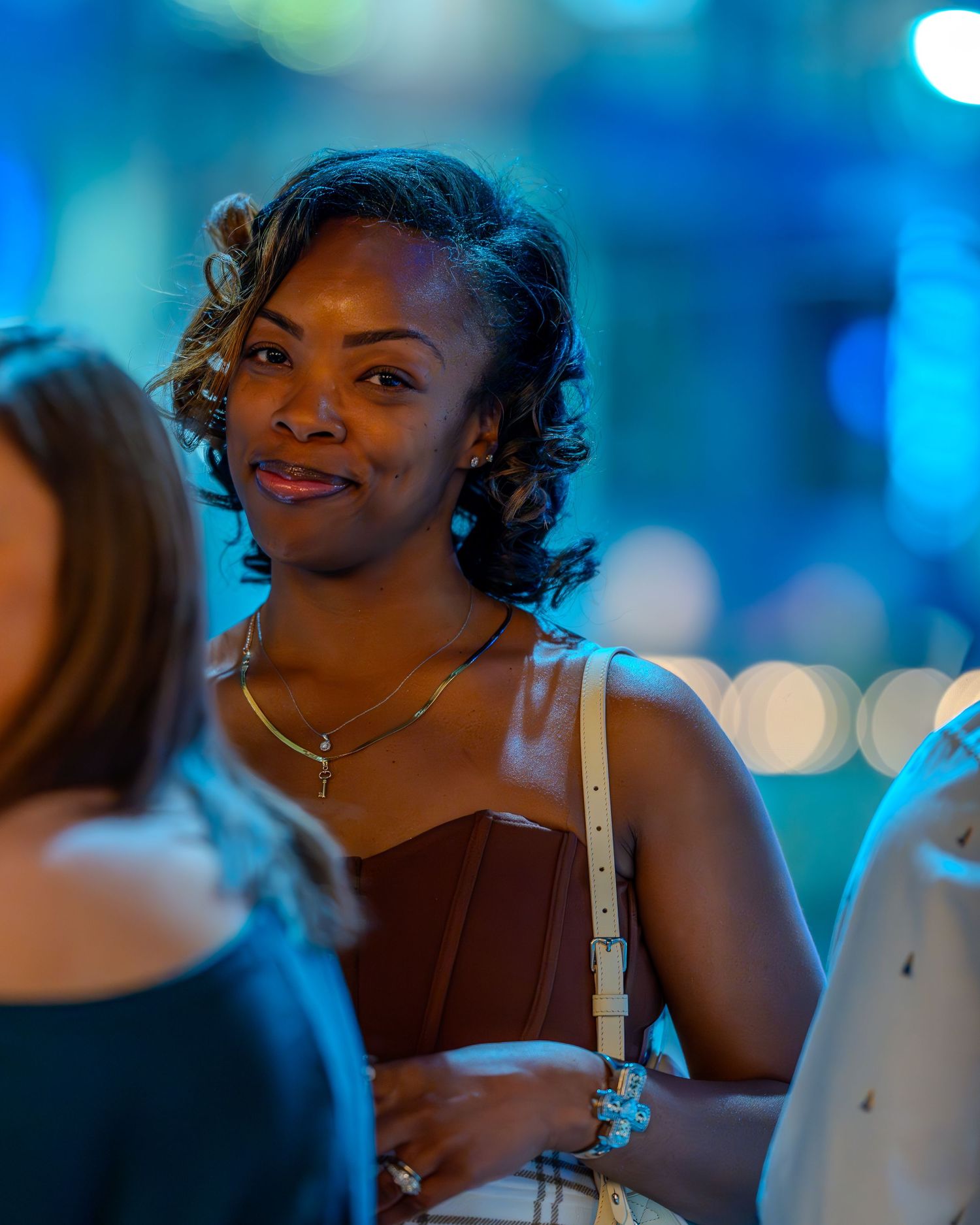 A smiling Black woman in a brown top and gold necklace stands outdoors at night with bokeh blue lights behind her.