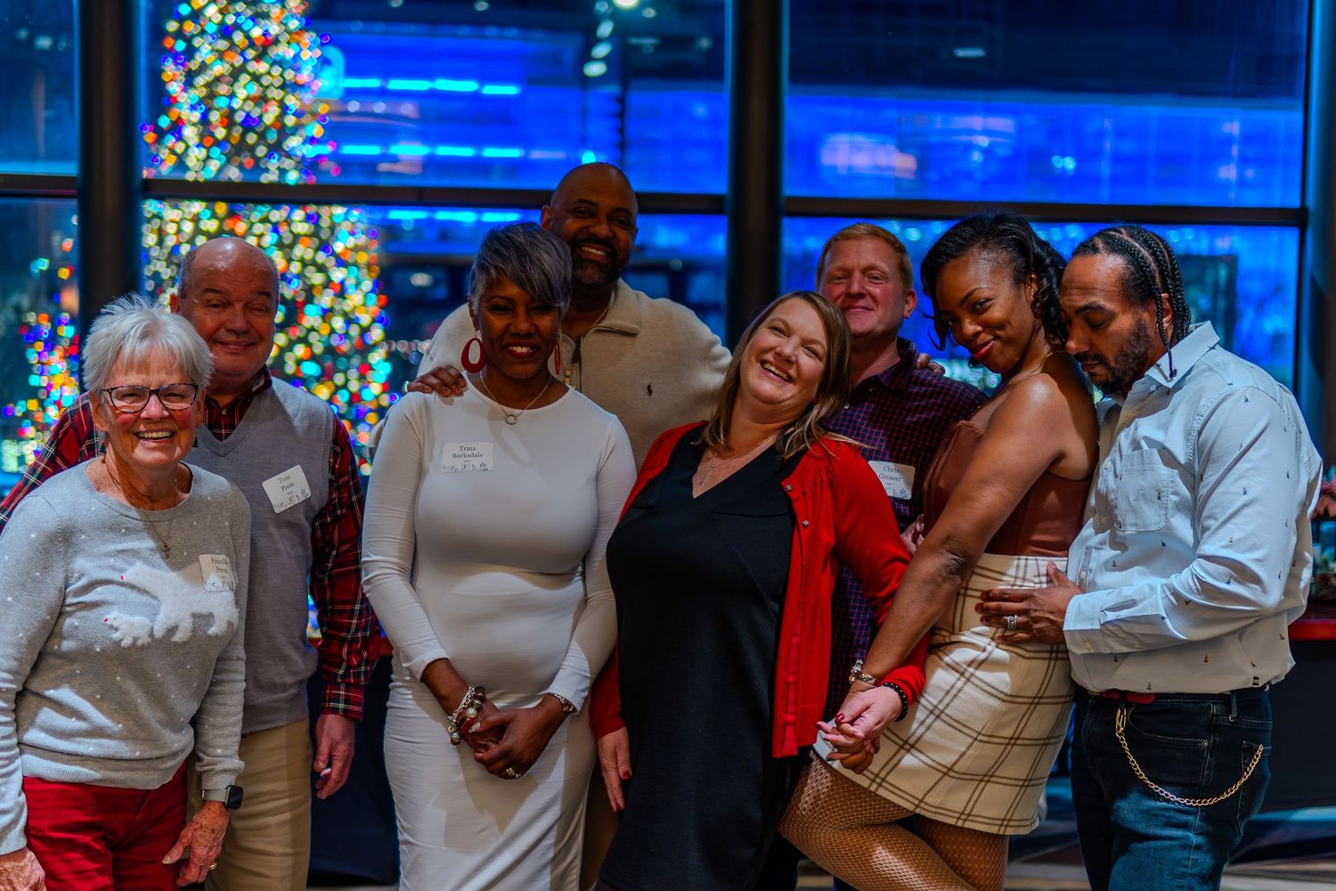 A diverse group of smiling friends posing together at a festive holiday party with a lit Christmas tree in the background.