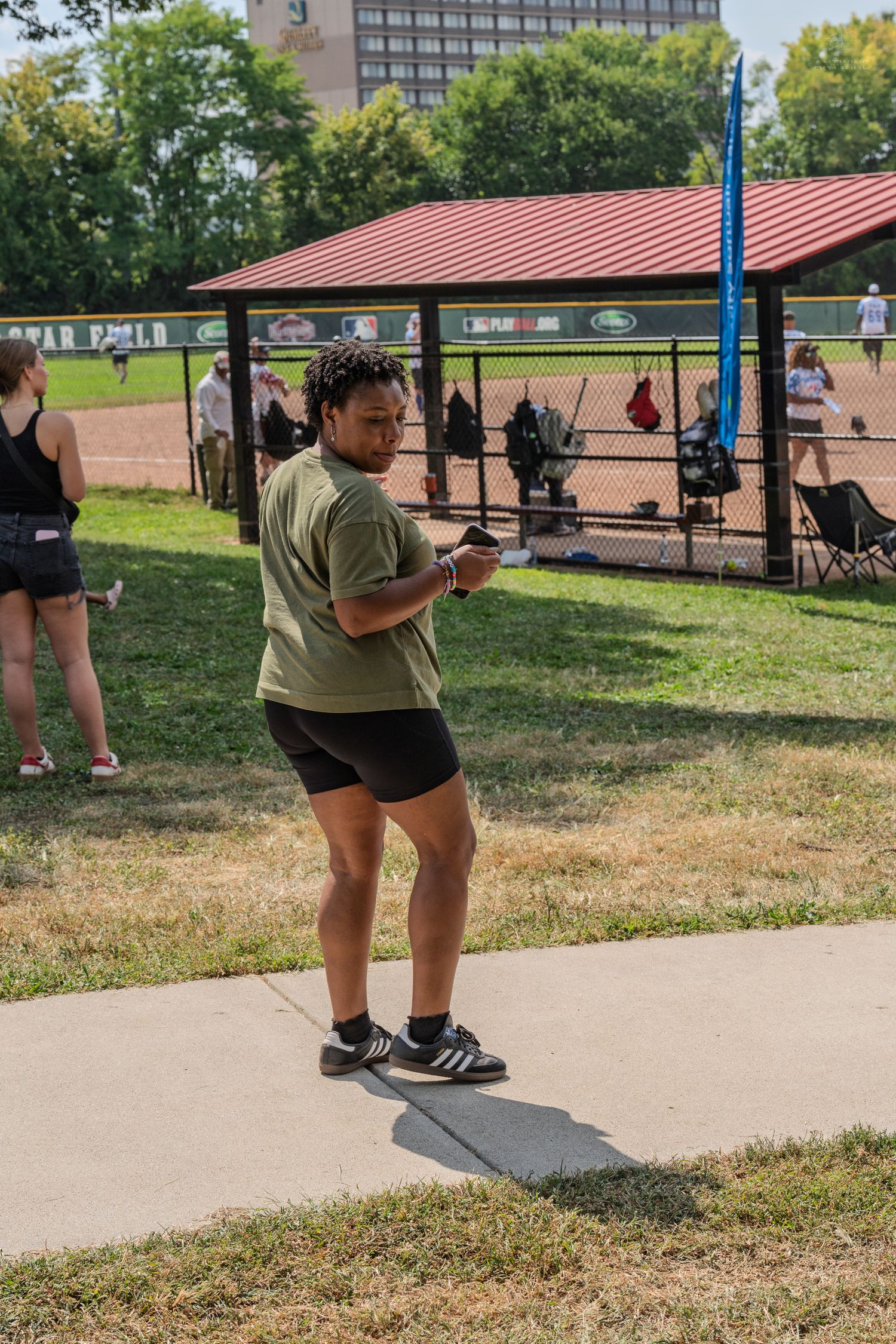 Woman in olive green shirt and black shorts checking her phone at an outdoor softball field on a sunny day.