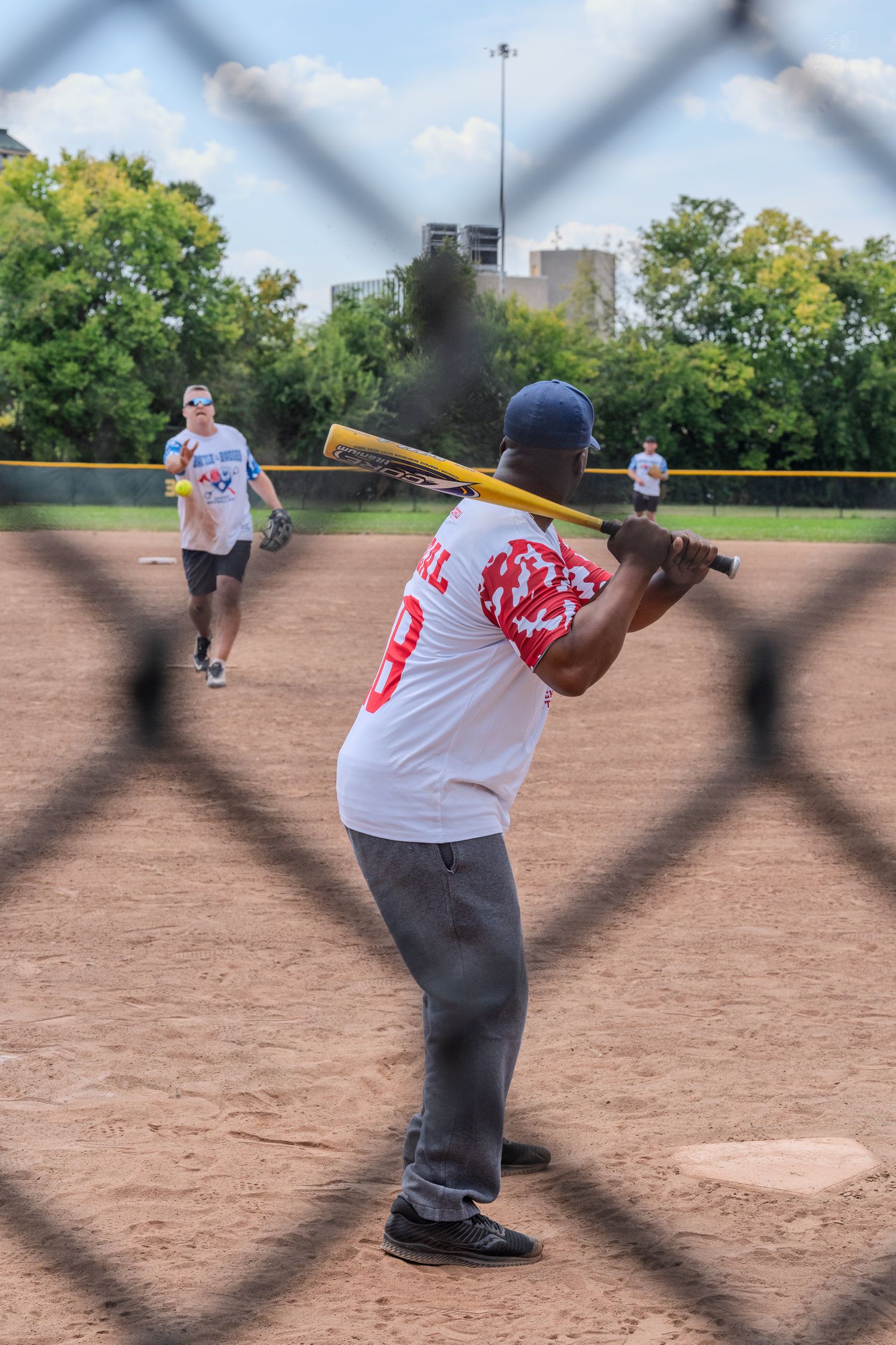 A batter in a red jersey swings at a pitch during an outdoor recreational softball game.