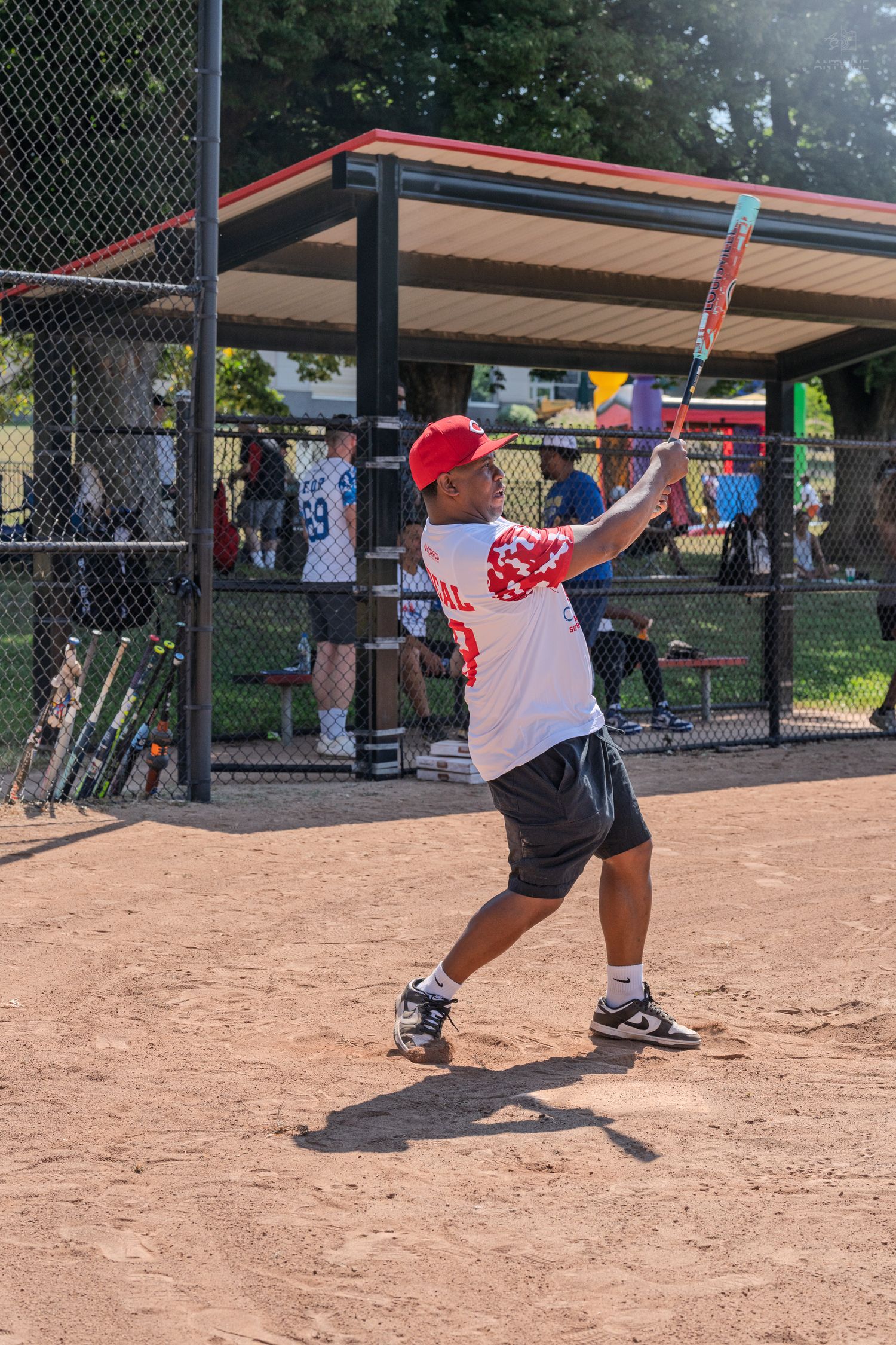 A batter in a red cap and white jersey swings a bat during a softball game on a dirt field.