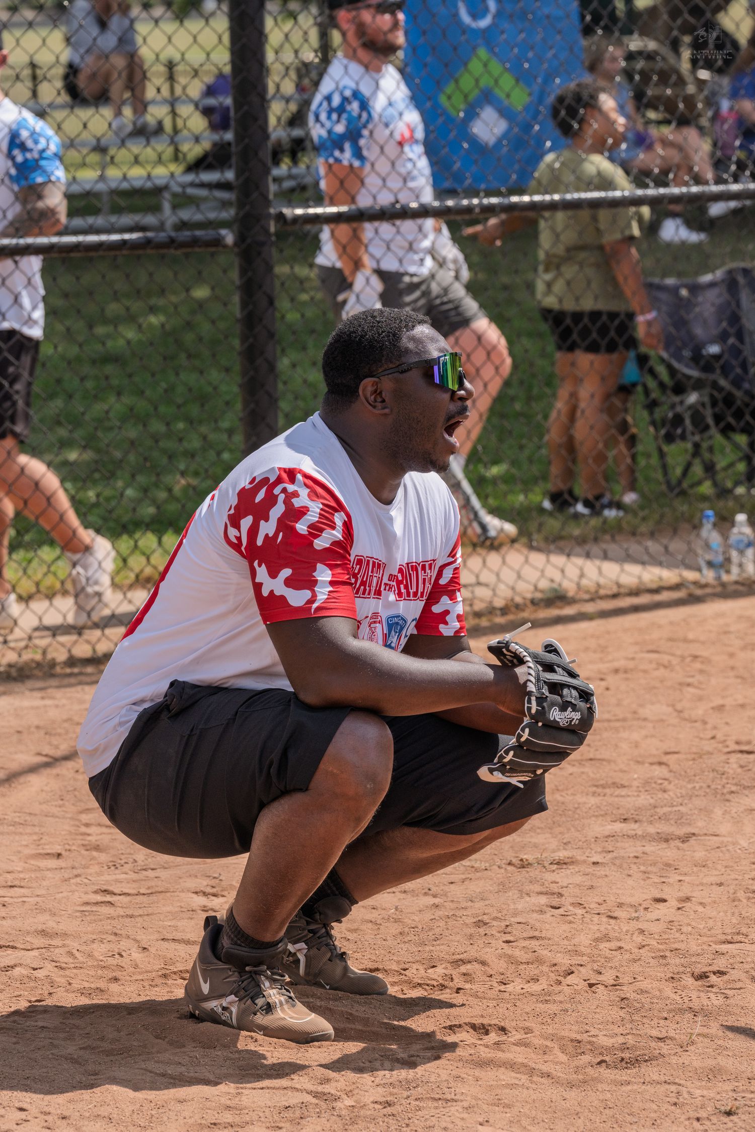 Male softball catcher crouching in position wearing glove and sunglasses during charity game.