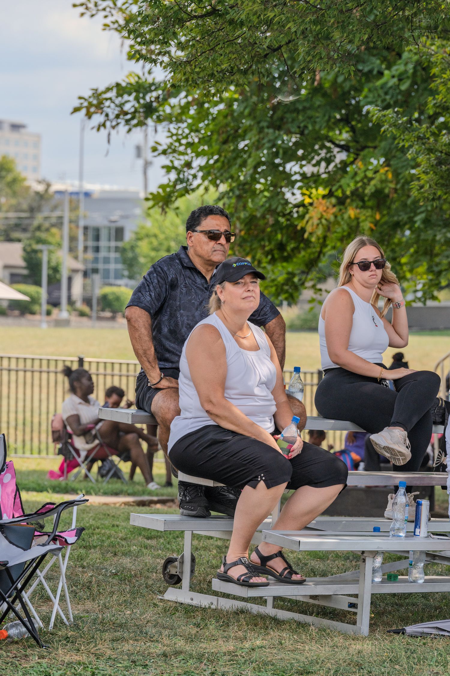 Three spectators sitting on bleachers watching an outdoor event on a sunny day in a park.