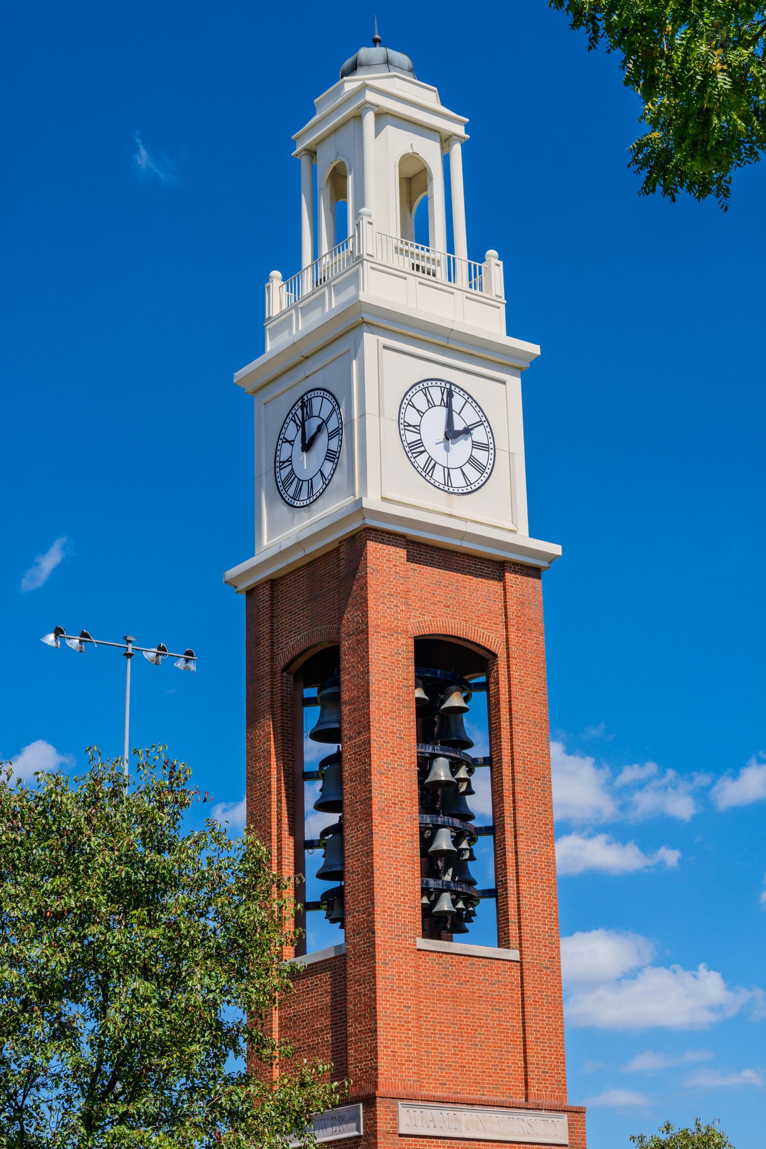 Brick clock tower with Roman numeral clock faces and bronze bells against a blue sky.