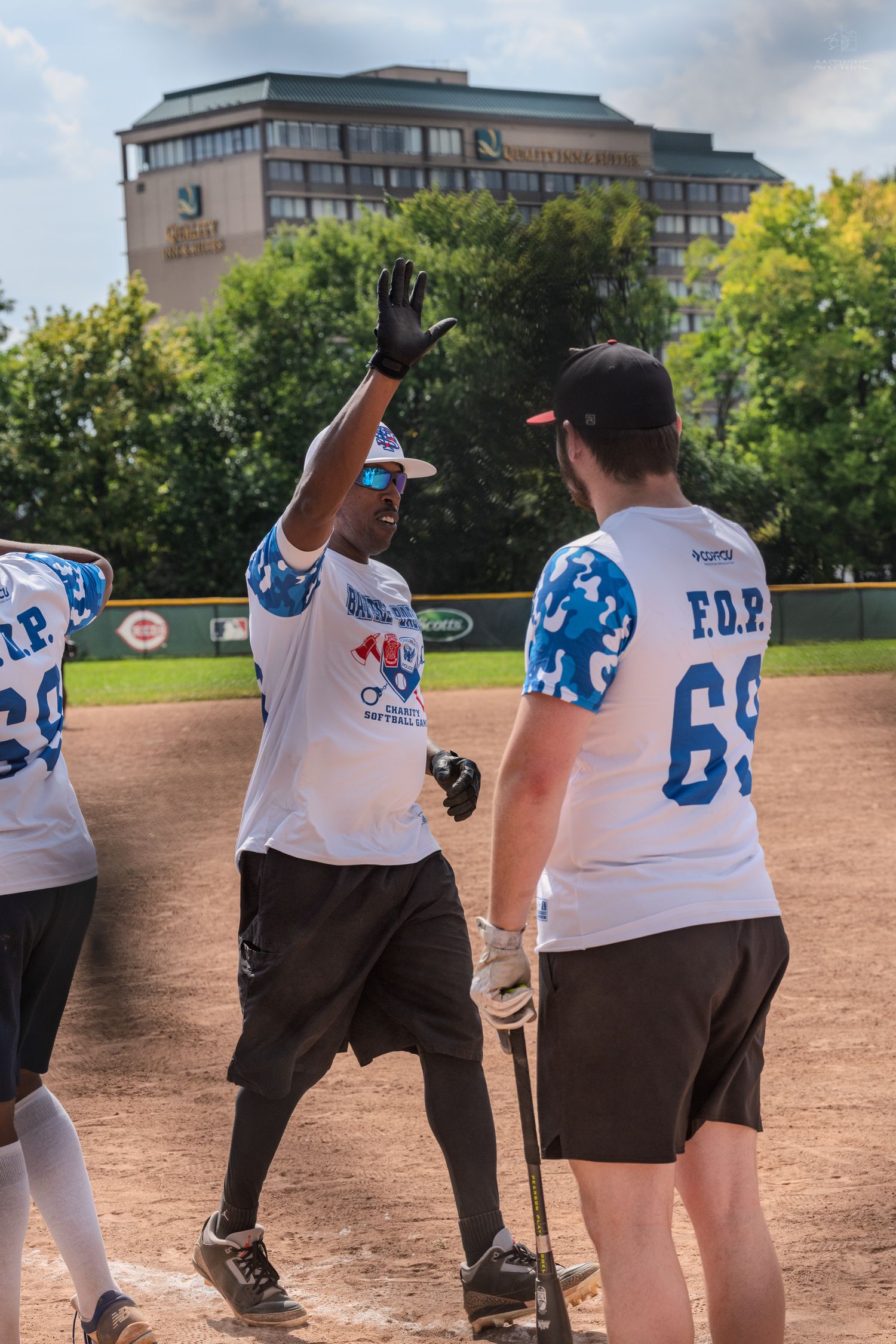 Two softball players in blue and white F.O.P. jerseys celebrate with a high five on the baseball diamond.