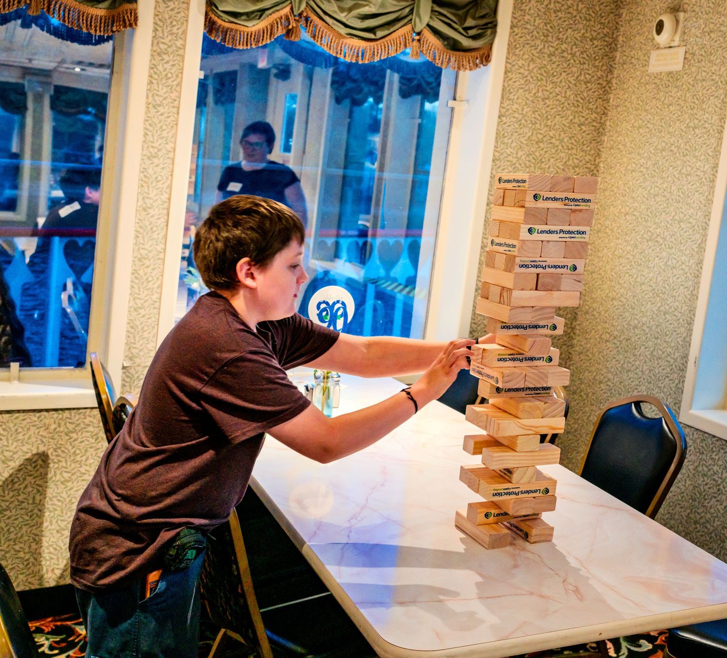 A boy carefully plays a giant Jenga tower game at a table indoors during a party event.