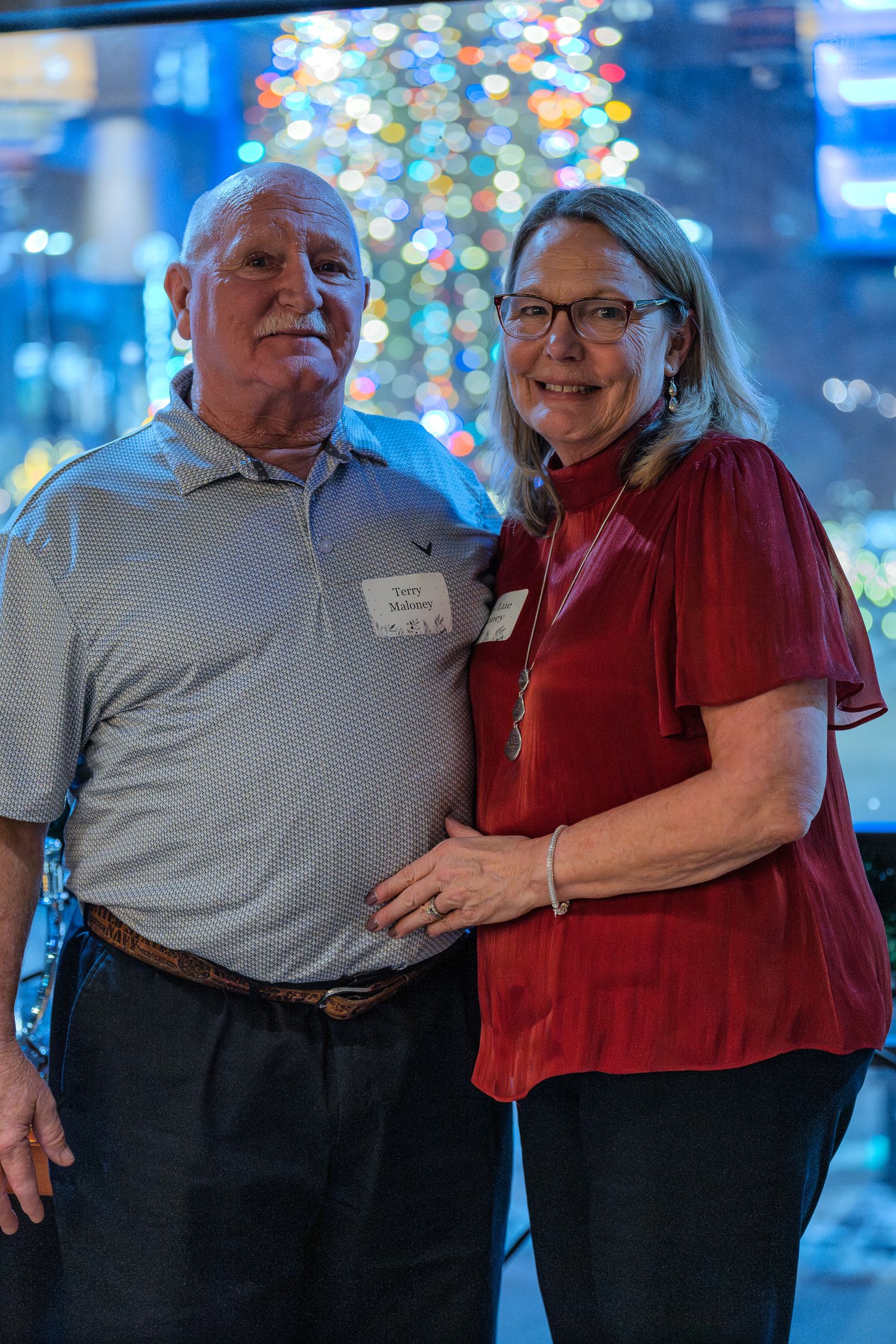 Smiling couple posing together at a festive holiday party with a decorated Christmas tree in the background.