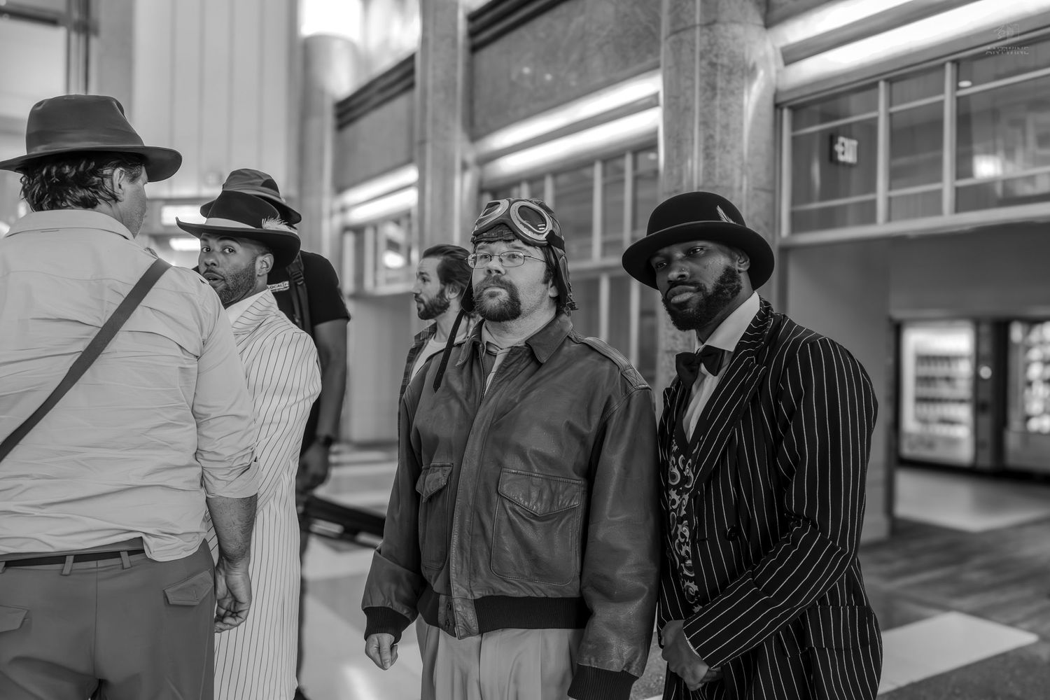 Black and white photo of men in vintage attire, including pinstripe suit and aviator jacket, gathered in a modern building.