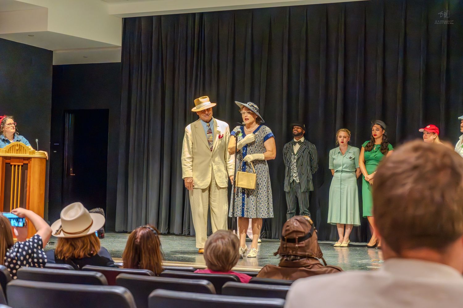 Actors in vintage 1940s costumes perform on stage during a theater production with audience seated in foreground.
