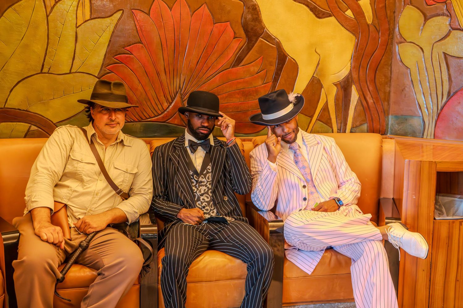 Three men in vintage 1920s-style suits and hats sitting on an orange sofa in front of an Art Deco mural.