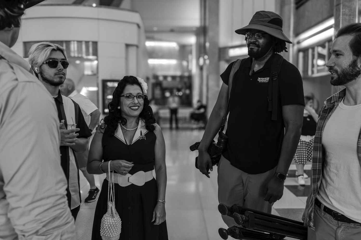 Black and white photo of a smiling woman socializing with a group of people in a large indoor venue.