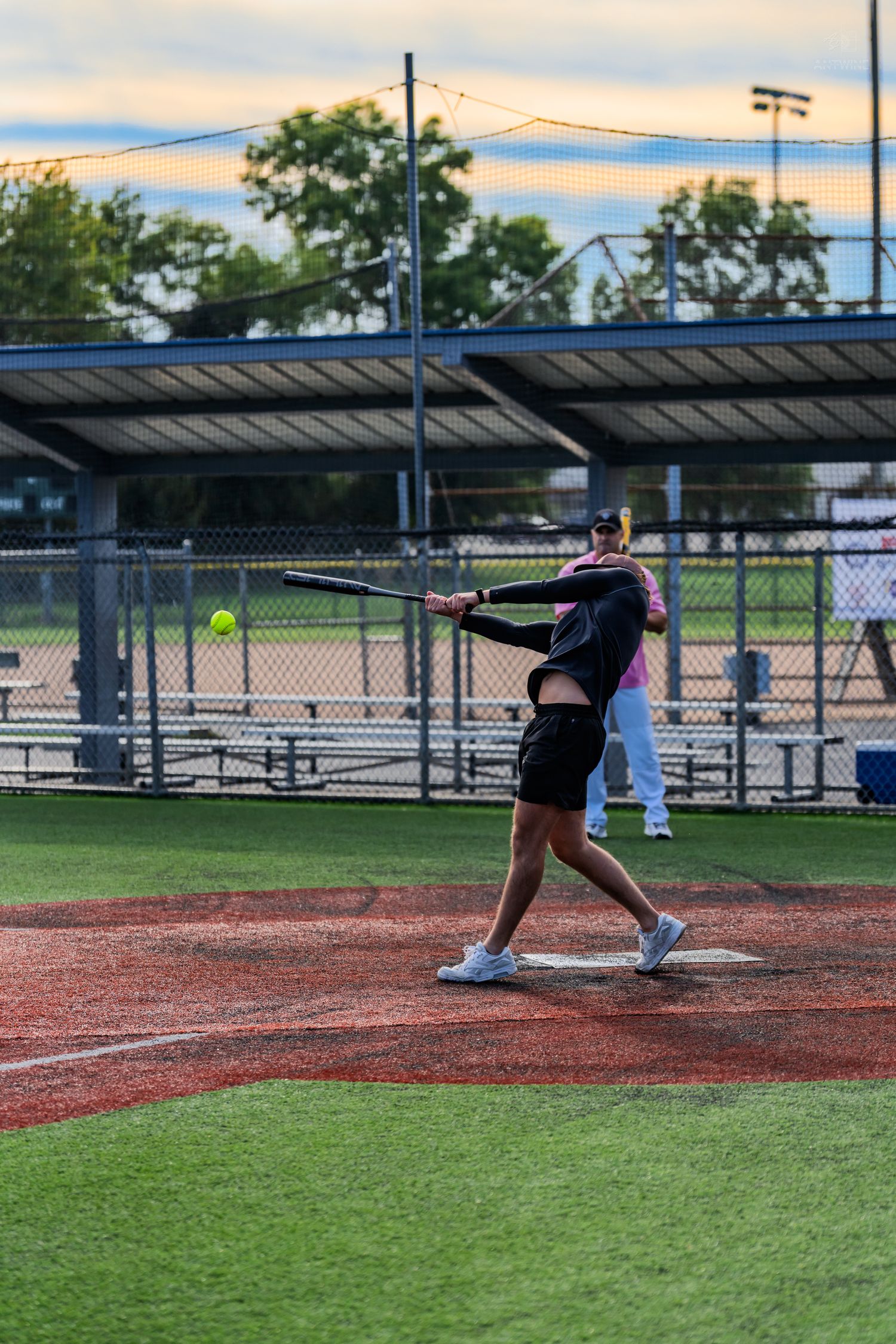 A softball player swings a bat at a yellow ball during an evening game on a red clay field.
