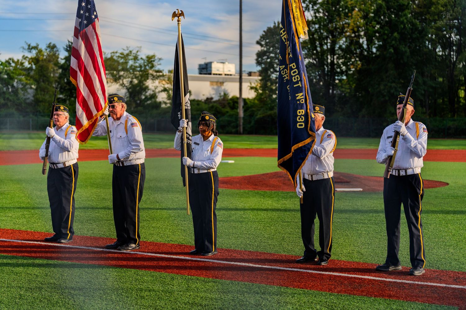 Military color guard presenting American and unit flags at outdoor baseball stadium ceremony.