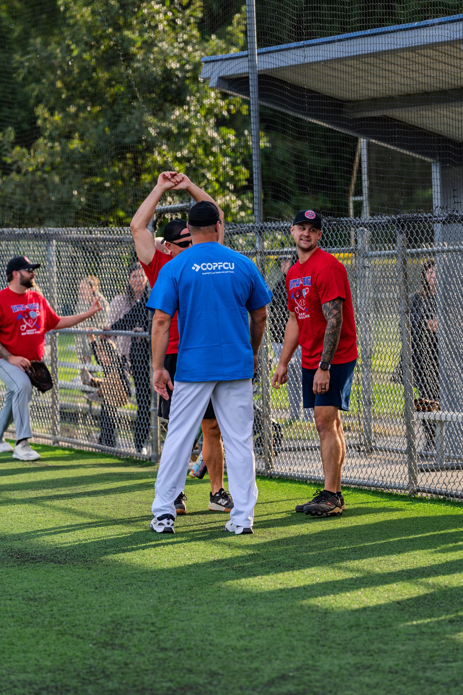 Baseball players in red and blue shirts interact near a chain-link fence dugout at an outdoor sports field.