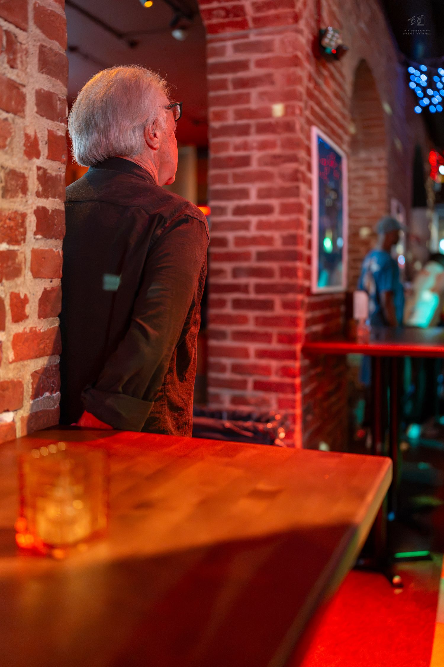 Elderly man with gray hair sitting alone at a bar with a drink, leaning against a red brick wall.