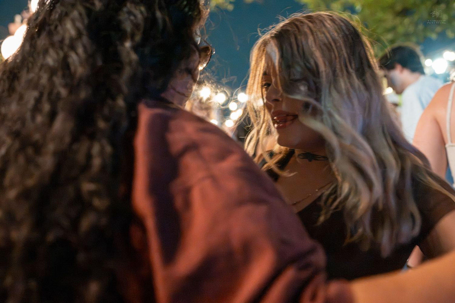 Two young women smiling and embracing at an outdoor evening party with string lights in the background.