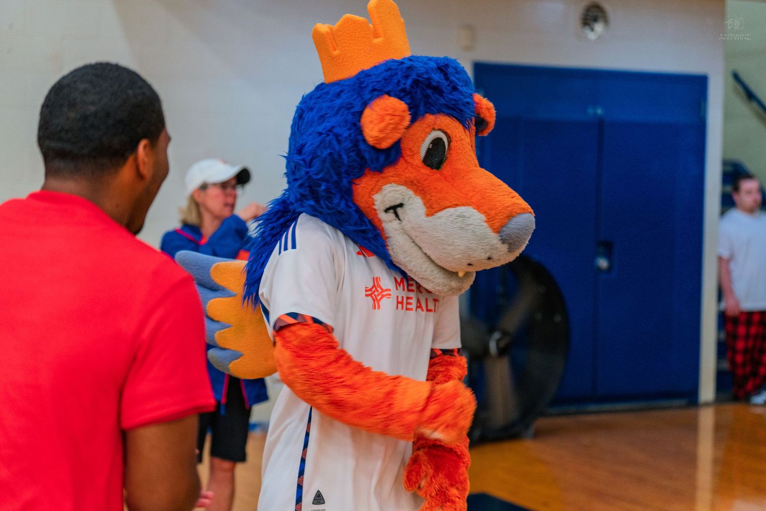 Orange and blue lion mascot in white jersey interacts with children on a gymnasium basketball court.