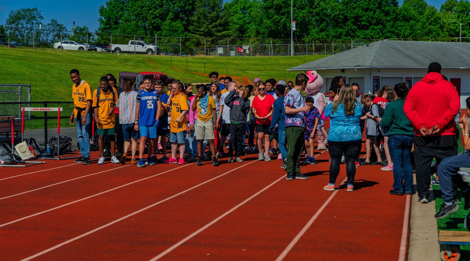 Students and participants gather on a red track at an outdoor school sports event on a sunny day.