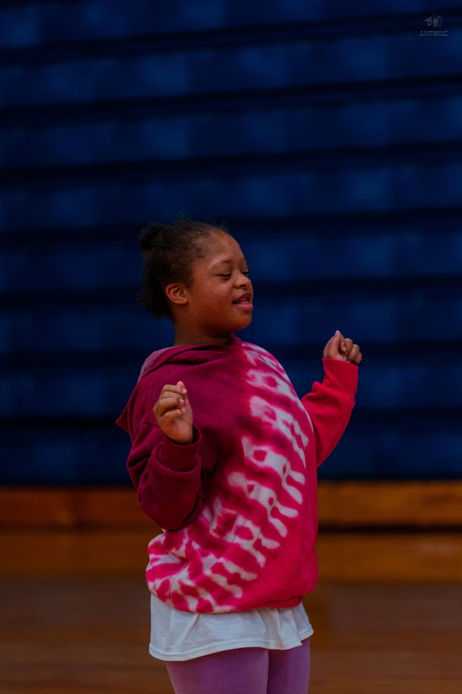 Young girl in pink tie-dye hoodie dancing joyfully on a gym floor with blue bleachers in background.
