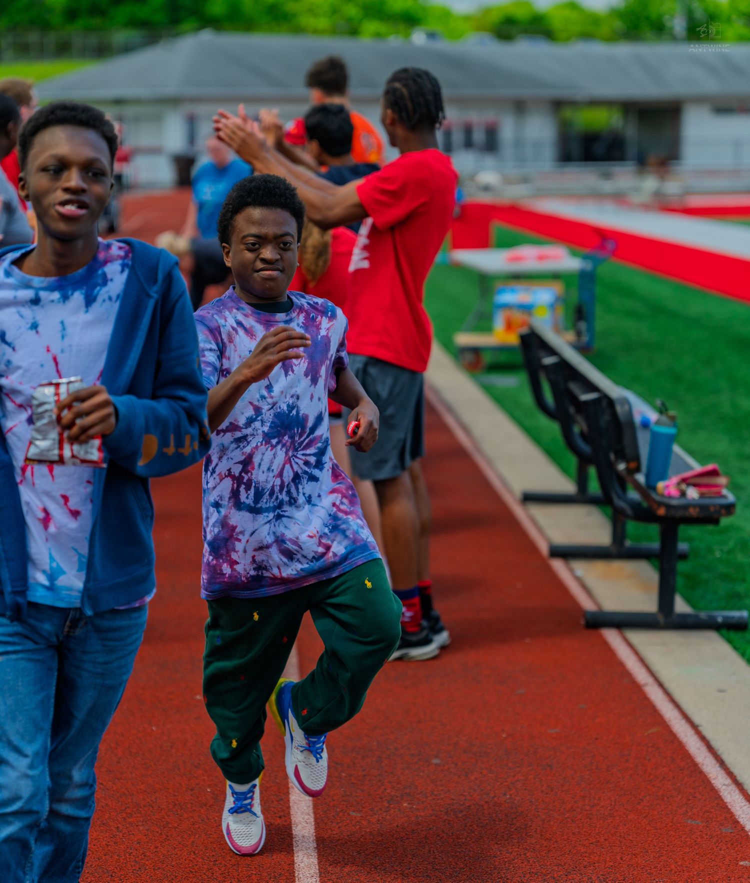 Two young athletes run along a red track while peers cheer and high-five them at a school track event.