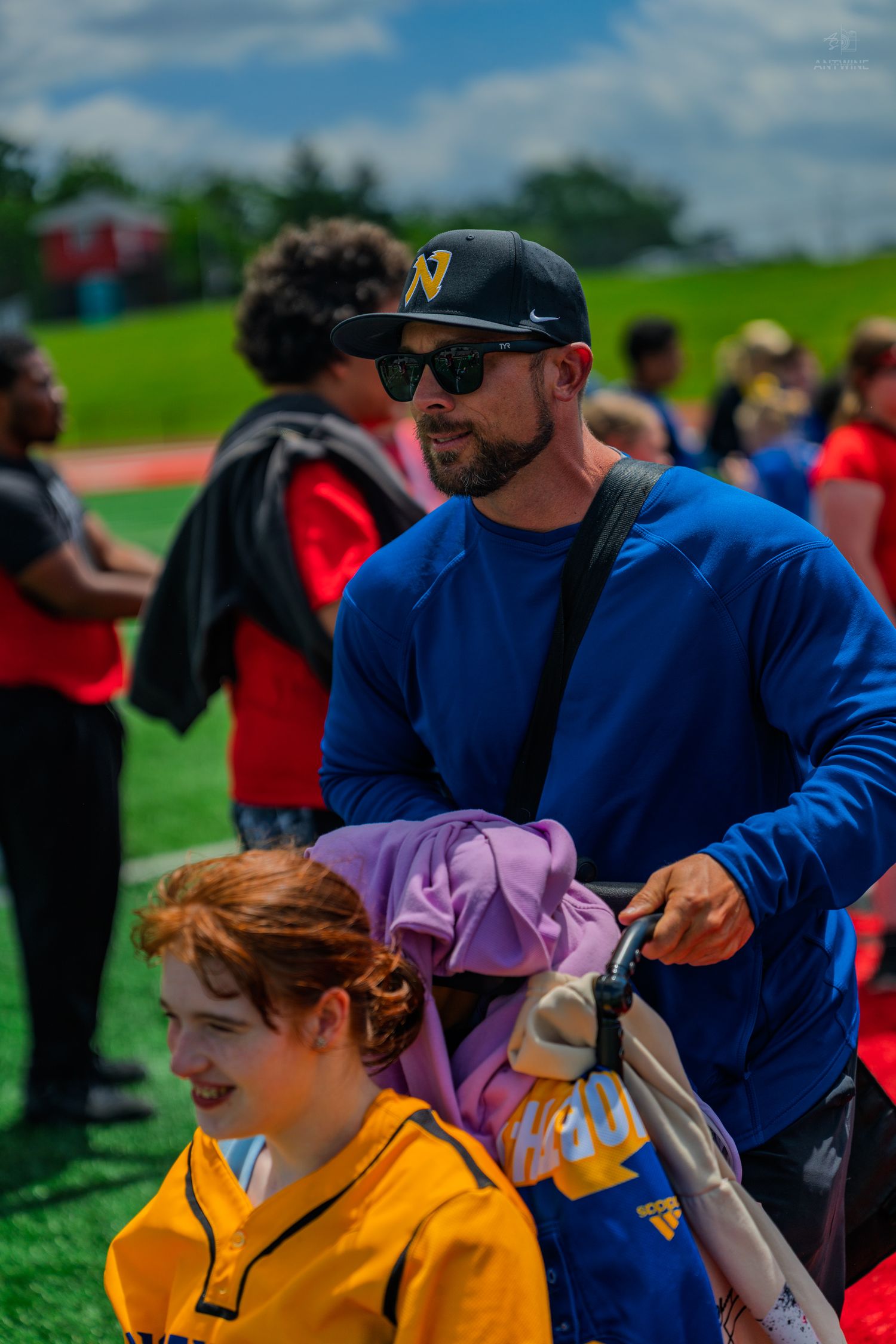 Man in blue shirt and cap pushes wheelchair at outdoor sports event with crowd in background.