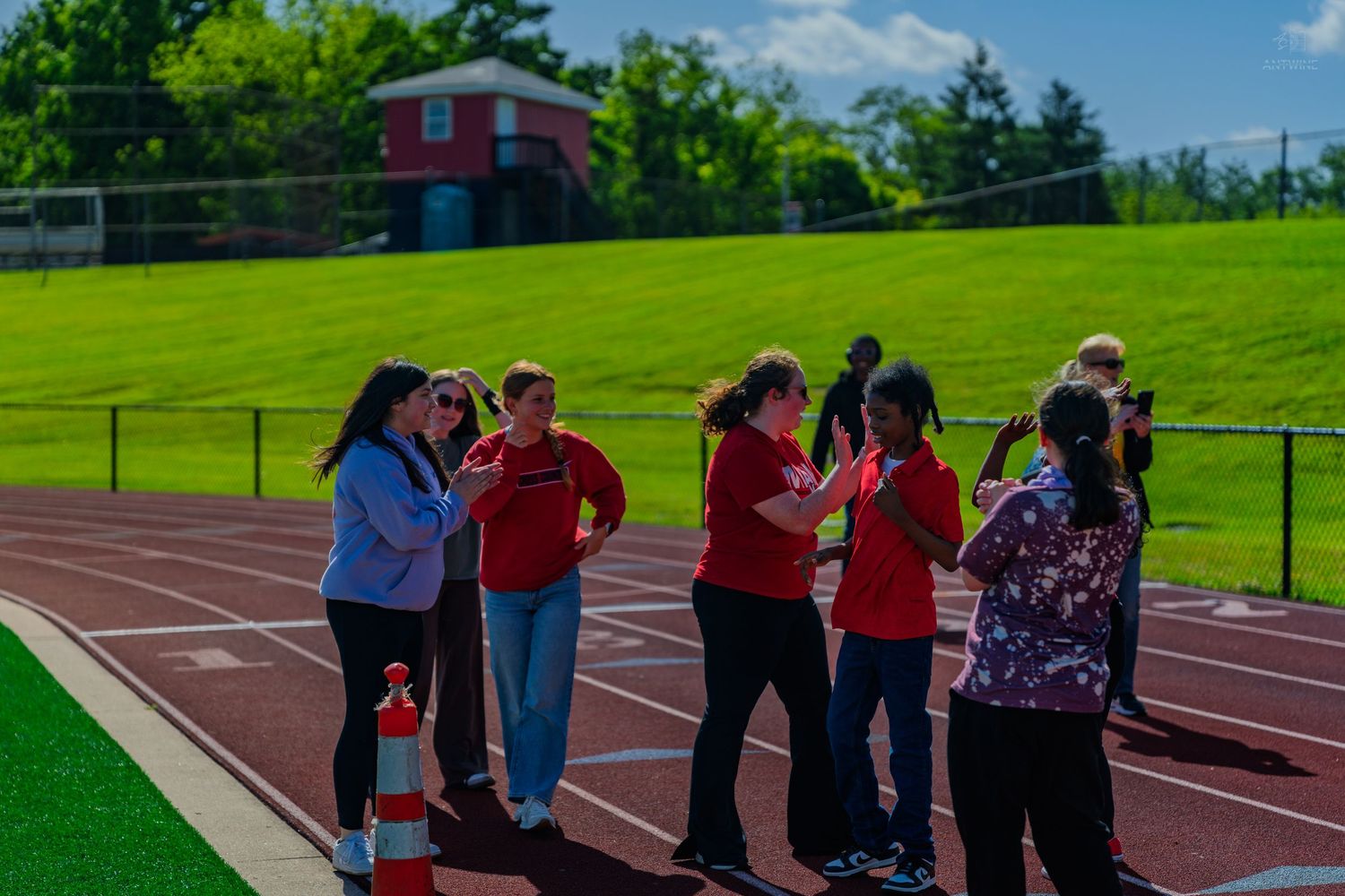 Group of people in red shirts gathered on a running track at an outdoor athletic field on a sunny day.