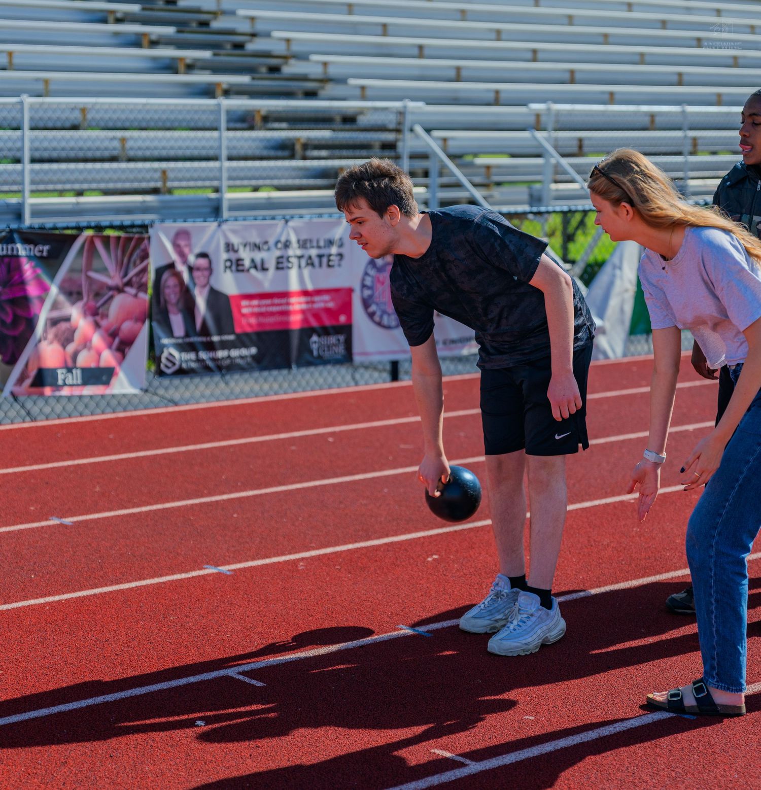 Two students play with a bowling ball on a red track at an outdoor stadium during a school event.