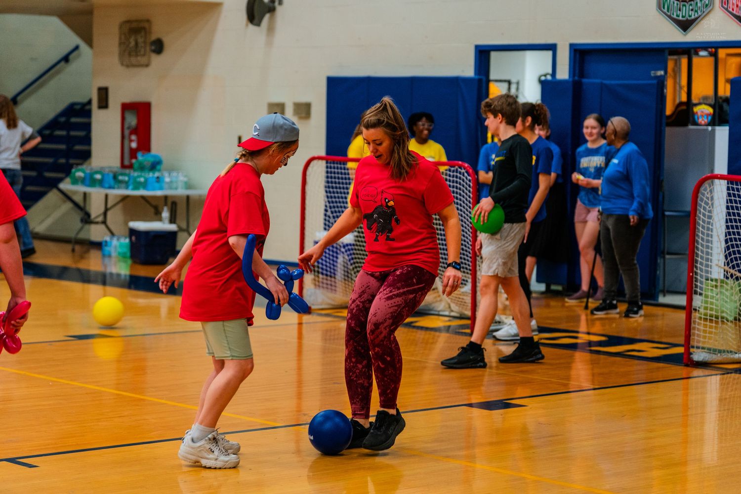 Students playing indoor soccer on a gymnasium court, with players in red shirts dribbling a ball toward a blue goal.