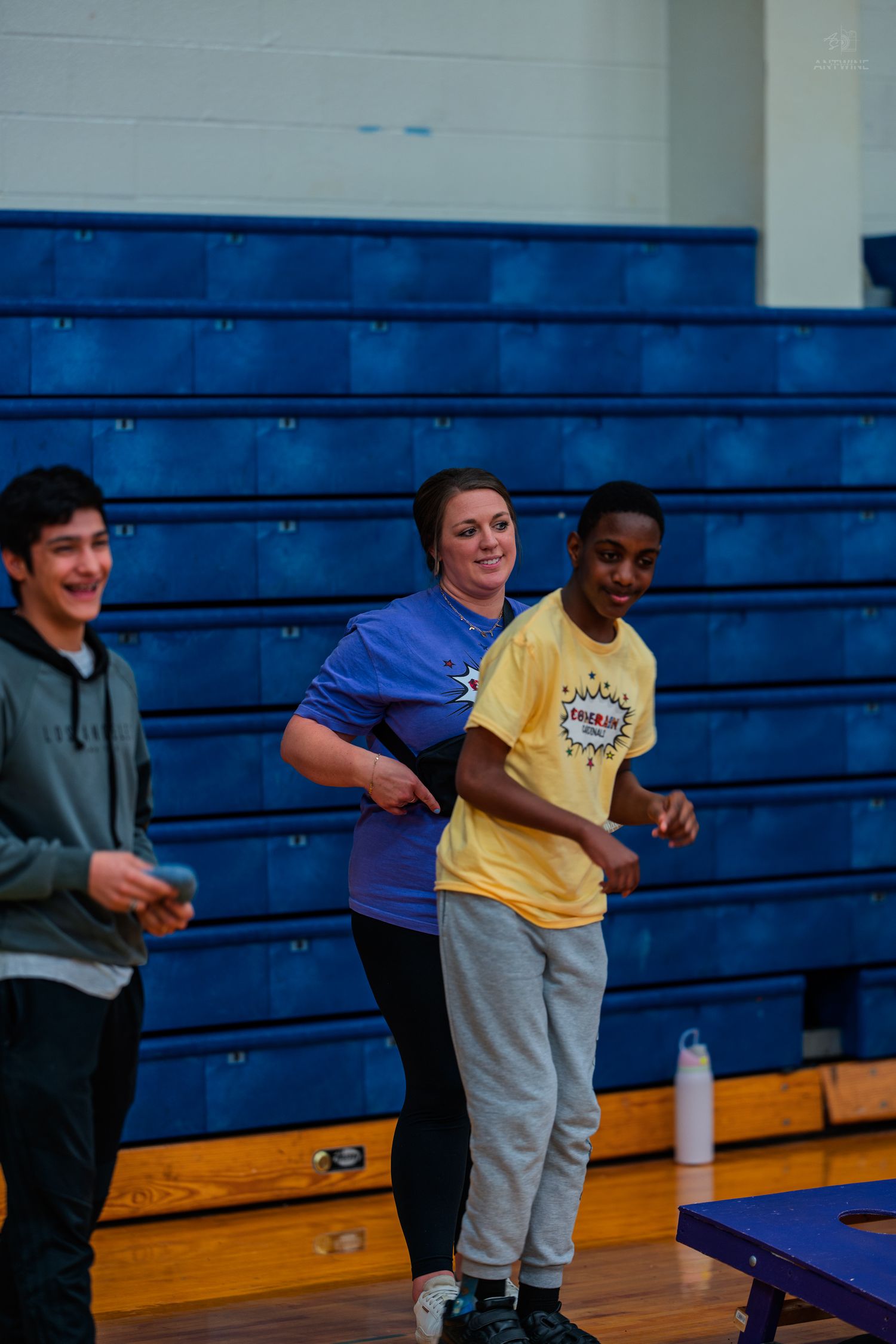 Students and teacher playing cornhole game in a school gymnasium with blue bleachers in the background.